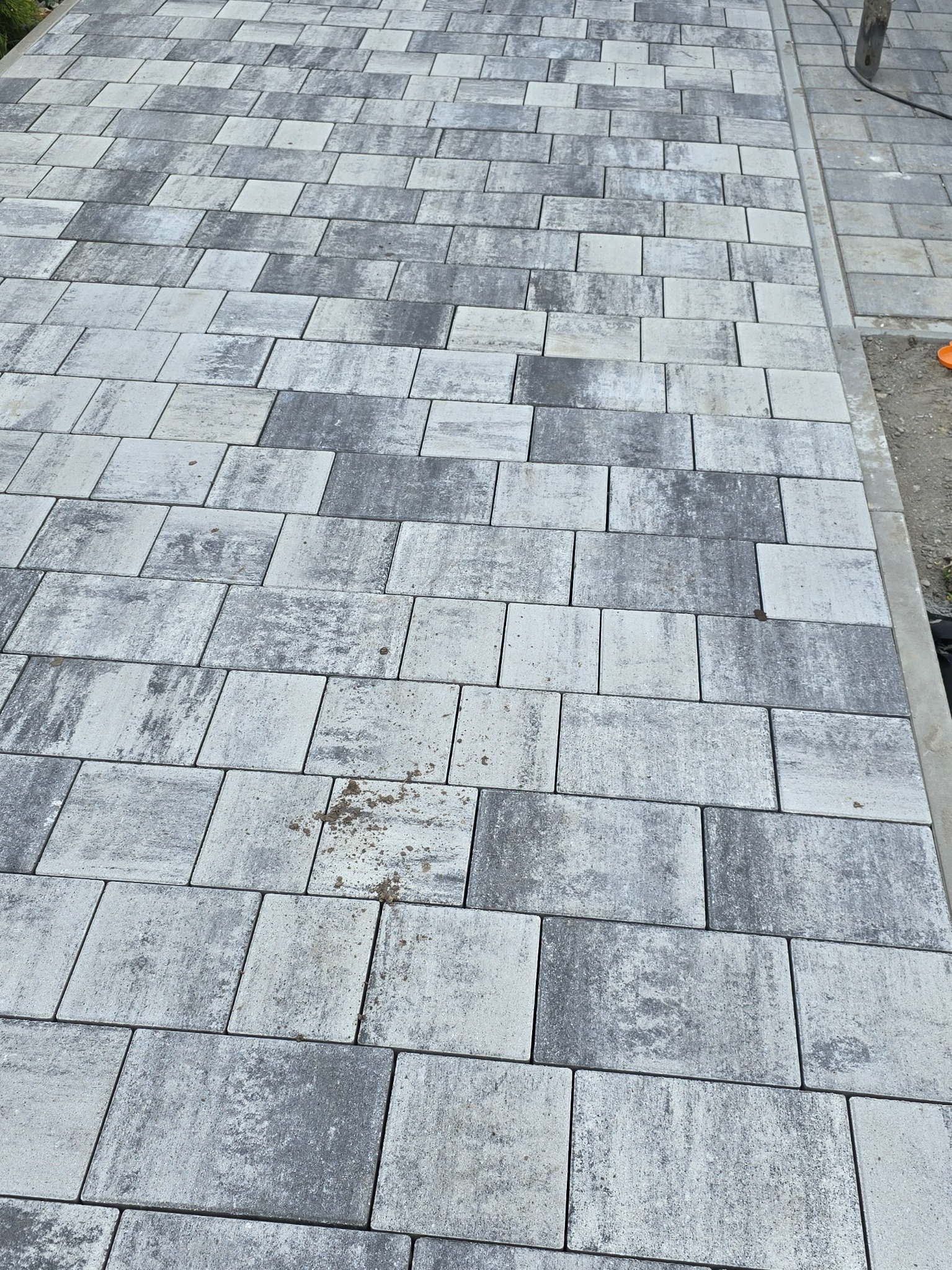 Ułożona kostka brukowa w odcieniach szarości, wzór prostokątny z delikatnymi zabrudzeniami w spoinach. Widok z góry, z fragmentem krawężnika po prawej.