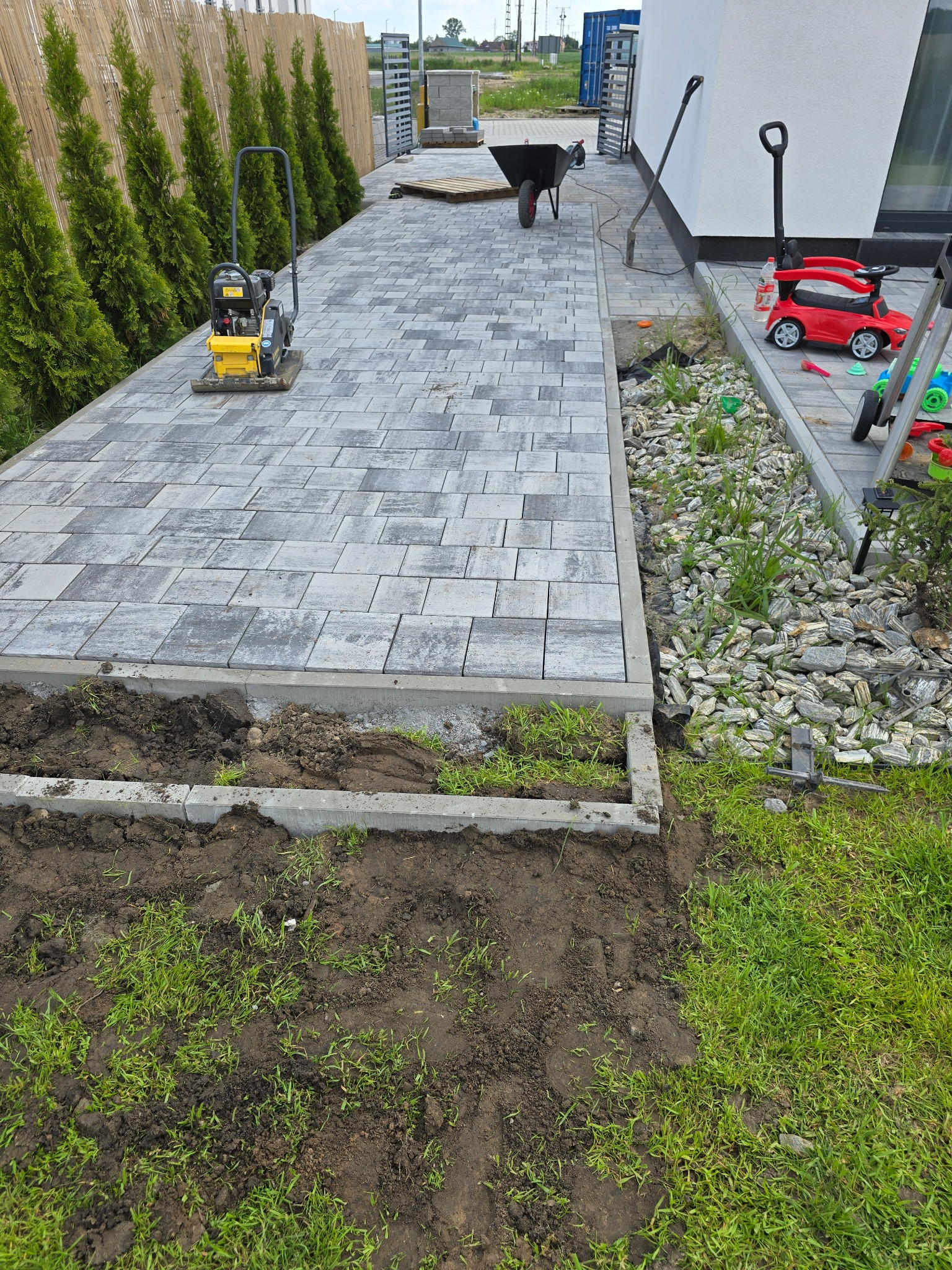 Fragment posesji w trakcie prac brukarskich: ułożona kostka, ubijarka, taczka, wykop pod obrzeża. W tle tuje i ogrodzenie panelowe.