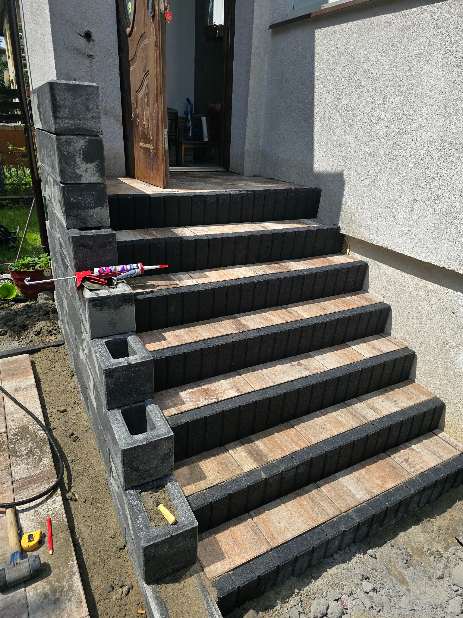 Schody wejściowe z kostki brukowej w trakcie montażu. Widoczne narzędzia: pistolet do kleju, młotek. Elementy kostki w kolorach szaro-czarnym i beżowo-brązowym.