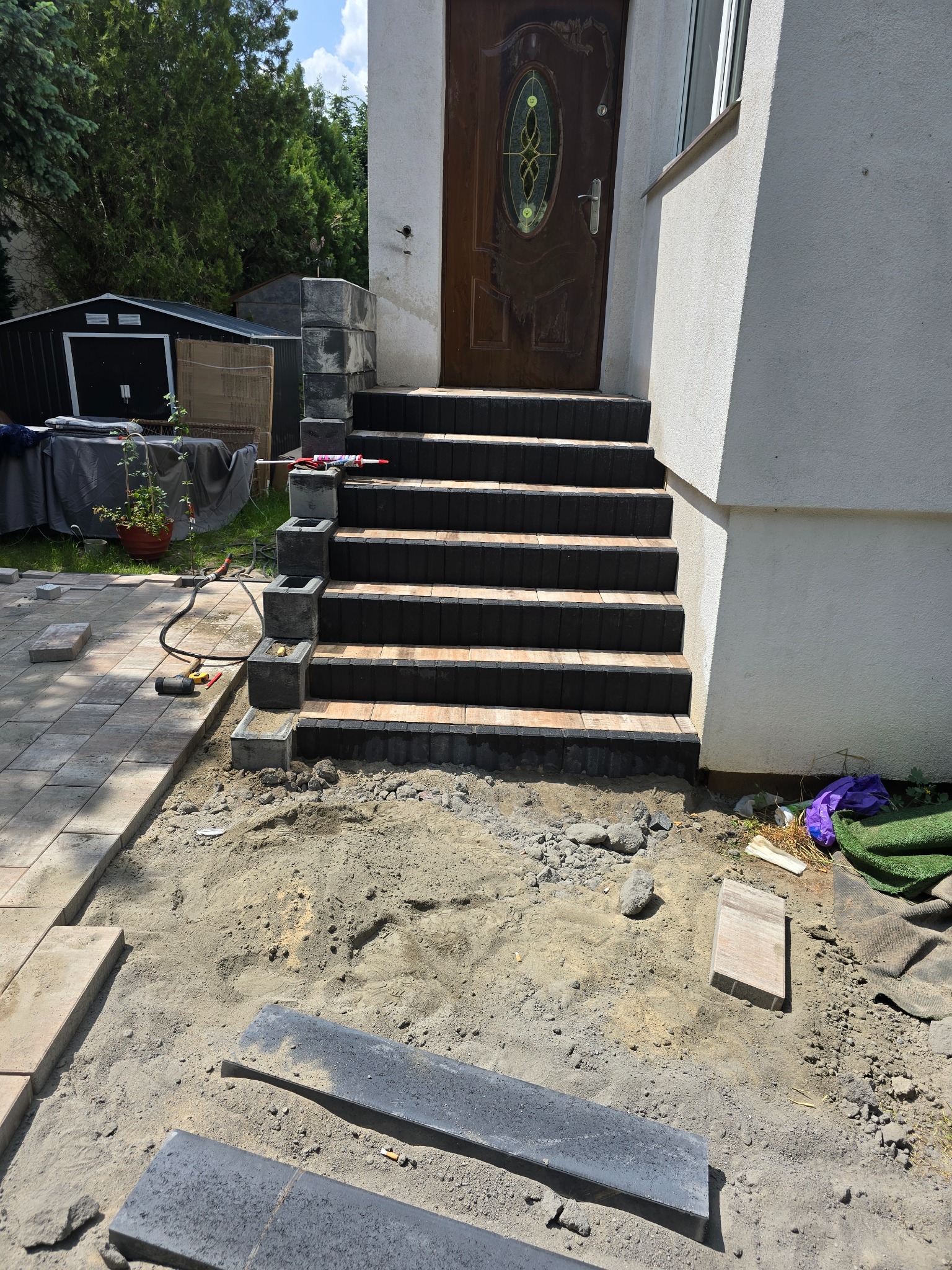 Schody wejściowe z kostki brukowej w trakcie budowy, z widocznymi narzędziami i materiałami budowlanymi, w tle zielony ogród i boczna ściana domu.