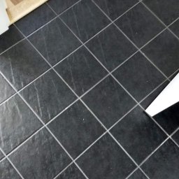 Prime-tech - Ciemnoszare, matowe płytki podłogowe ułożone w karo, z jasną fugą, widoczne przejście do drewnianej podłogi i fragment buta w rogu.