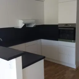 Nowoczesna kuchnia z białymi szafkami i czarnym blatem, widoczny zlewozmywak, okap i piekarnik, podłoga z paneli.