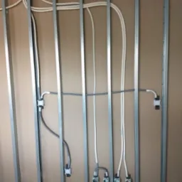 Stelaż z profili stalowych przytwierdzony do ściany z płyty karton-gips, z poprowadzonymi przewodami elektrycznymi i puszkami instalacyjnymi, widoczne złącza i kable w kolorach białym i szarym.