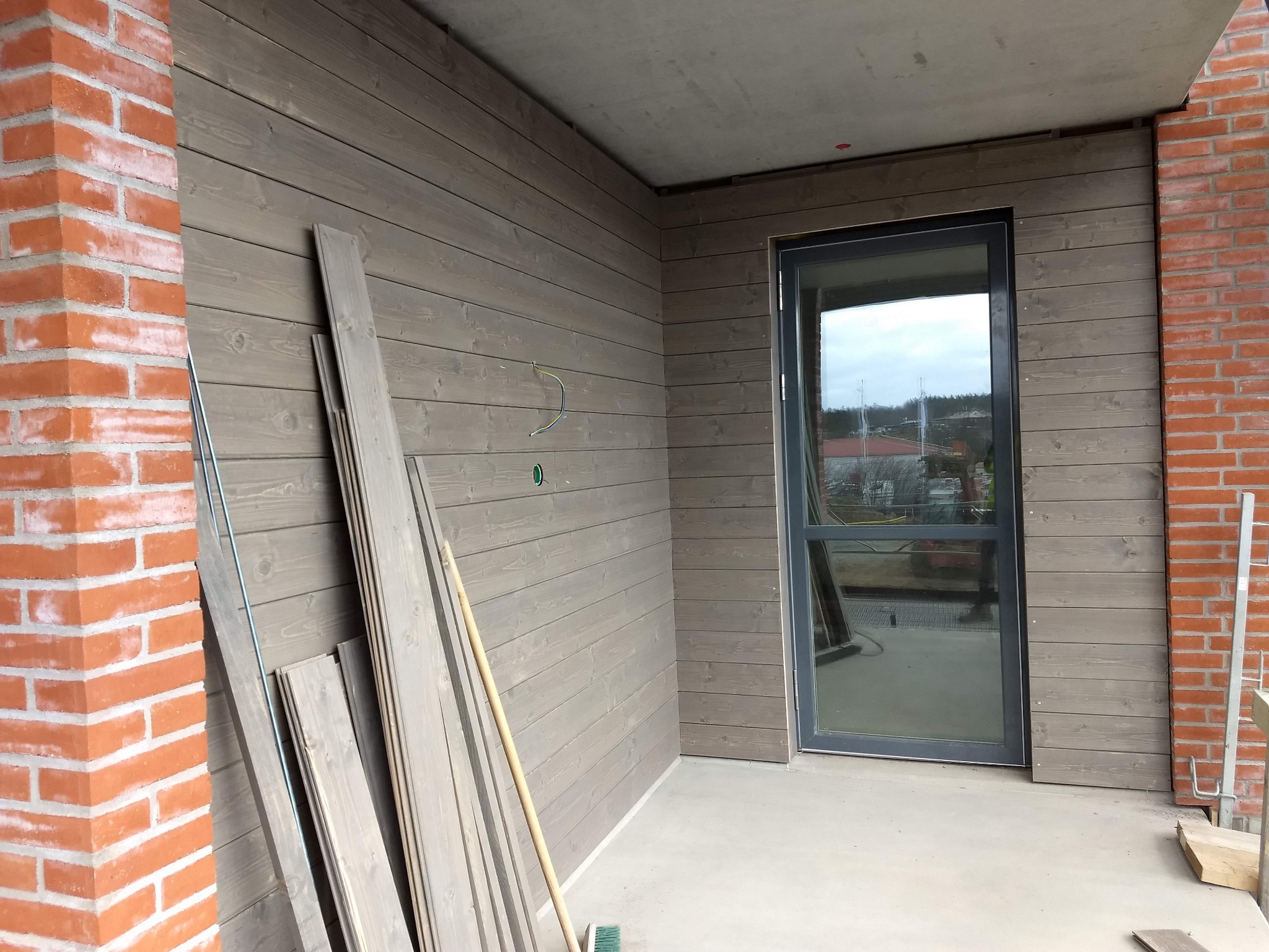 Montaż drewnianej boazerii na ścianie zewnętrznej budynku, widoczne deski przed montażem, ceglany narożnik i zamontowane drzwi z czarną ramą.