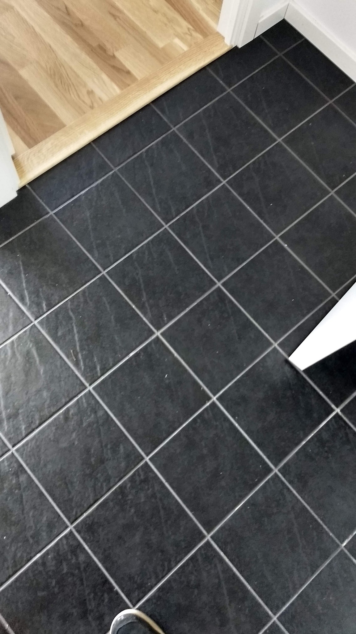 Ciemnoszare, matowe płytki podłogowe ułożone w karo, z jasną fugą, widoczne przejście do drewnianej podłogi i fragment buta w rogu.