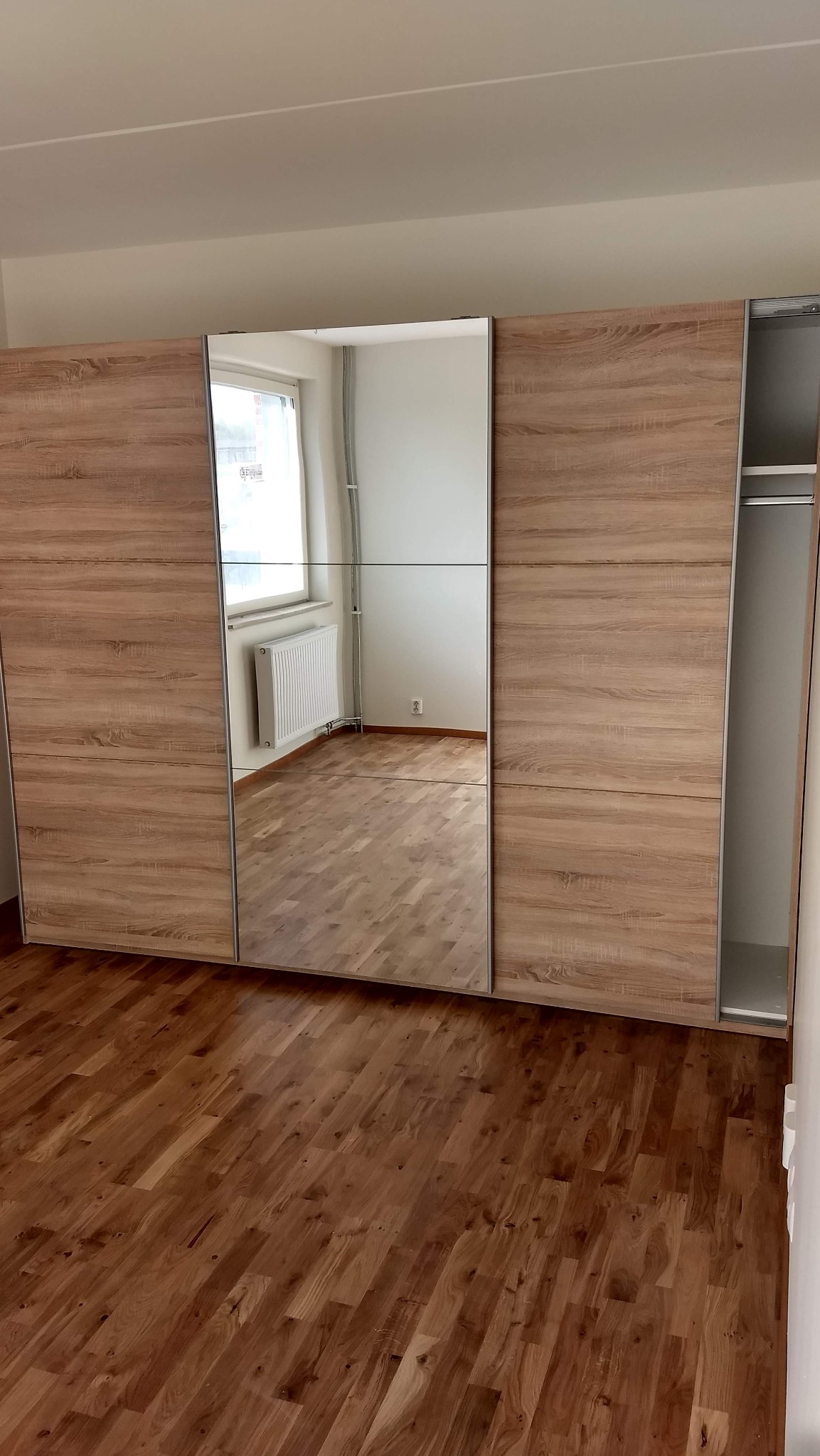 Nowoczesna szafa przesuwna z lustrzanymi drzwiami w jasnym pokoju z drewnianą podłogą, widoczny grzejnik i puste wnętrze szafy.