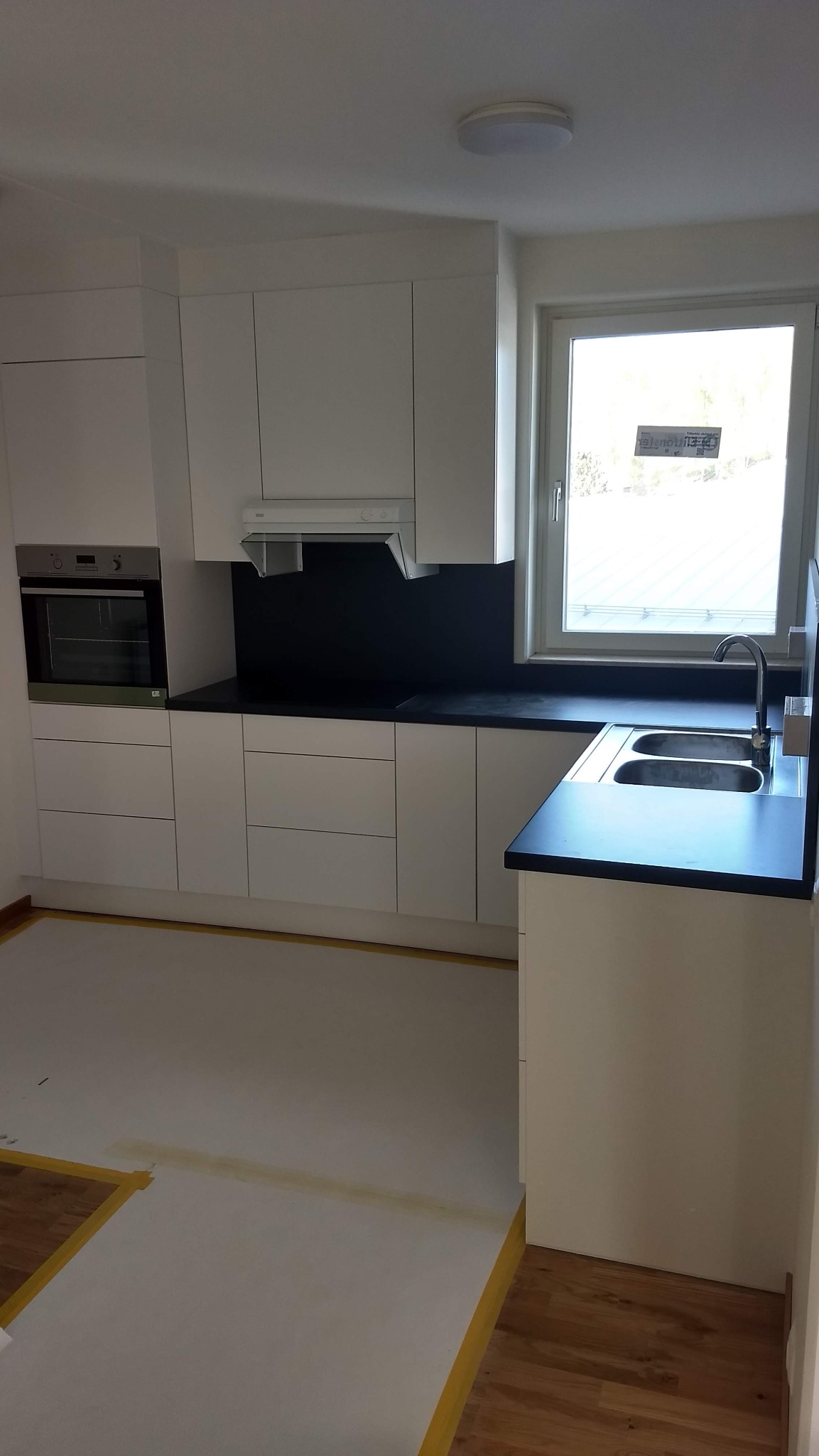 Wnętrze kuchni z białymi szafkami i czarnym blatem, widoczne zabezpieczenie podłogi taśmą malarską, okno z widokiem na zewnątrz.