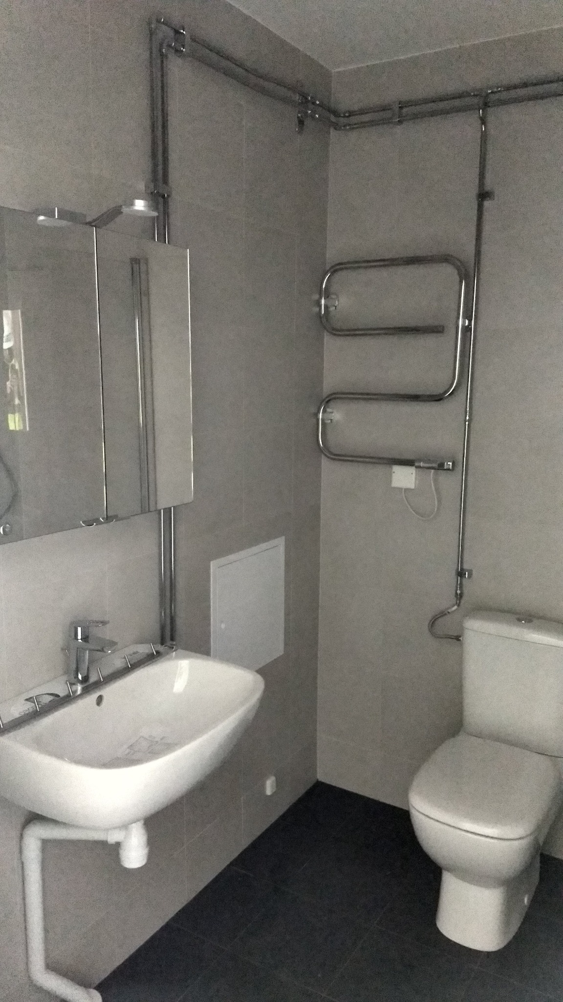 Wykończona łazienka z szarymi płytkami, białą umywalką, toaletą i grzejnikiem drabinkowym. Widoczne rury instalacji wodnej i kanalizacyjnej.