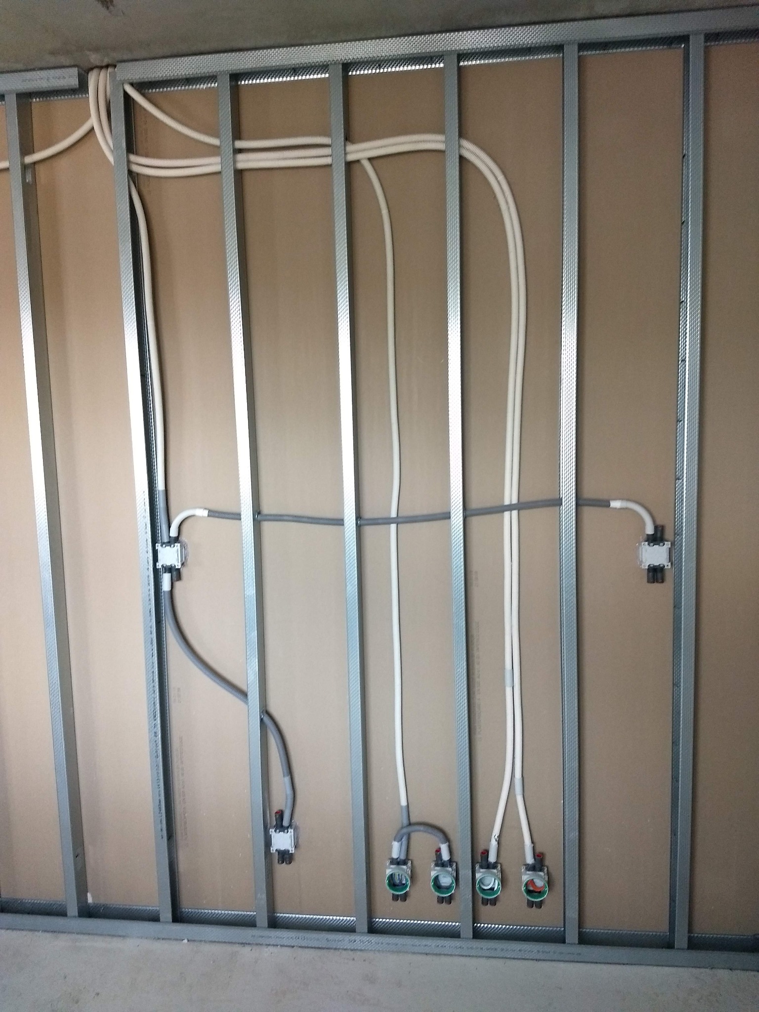 Stelaż z profili stalowych przytwierdzony do ściany z płyty karton-gips, z poprowadzonymi przewodami elektrycznymi i puszkami instalacyjnymi, widoczne złącza i kable w kolorach białym i szarym.