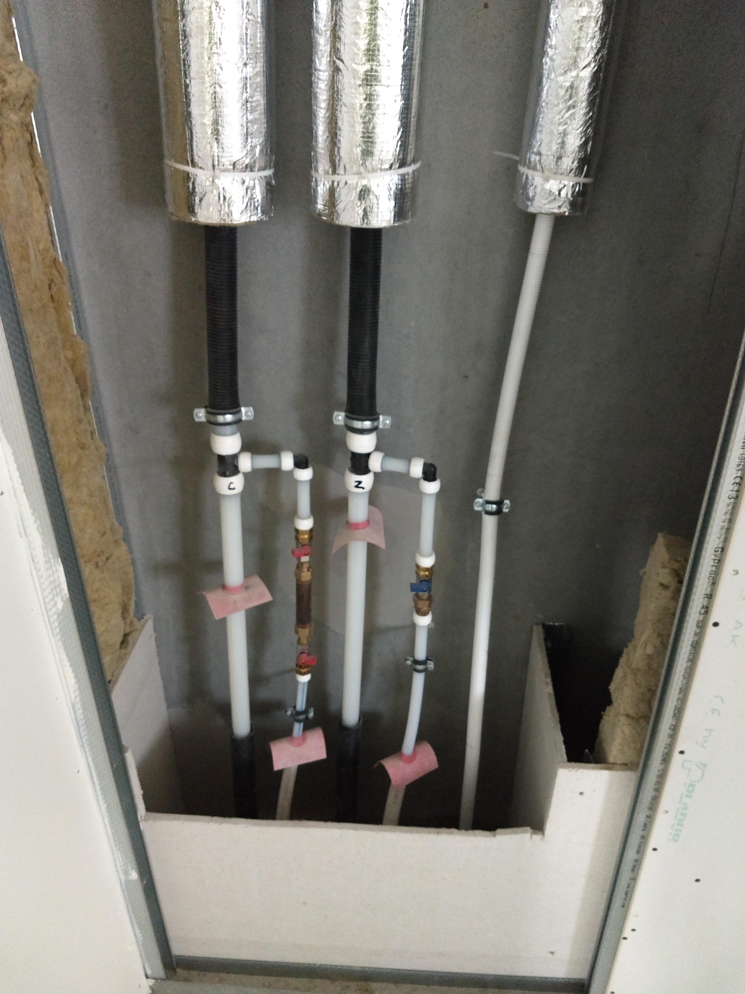 Instalacja rur grzewczych i wodnych w przestrzeni pod sufitem podwieszanym z płyty karton-gips, widoczne izolowane rury pionowe, białe rury plastikowe z zaworami, oraz fragment wełny mineralnej.