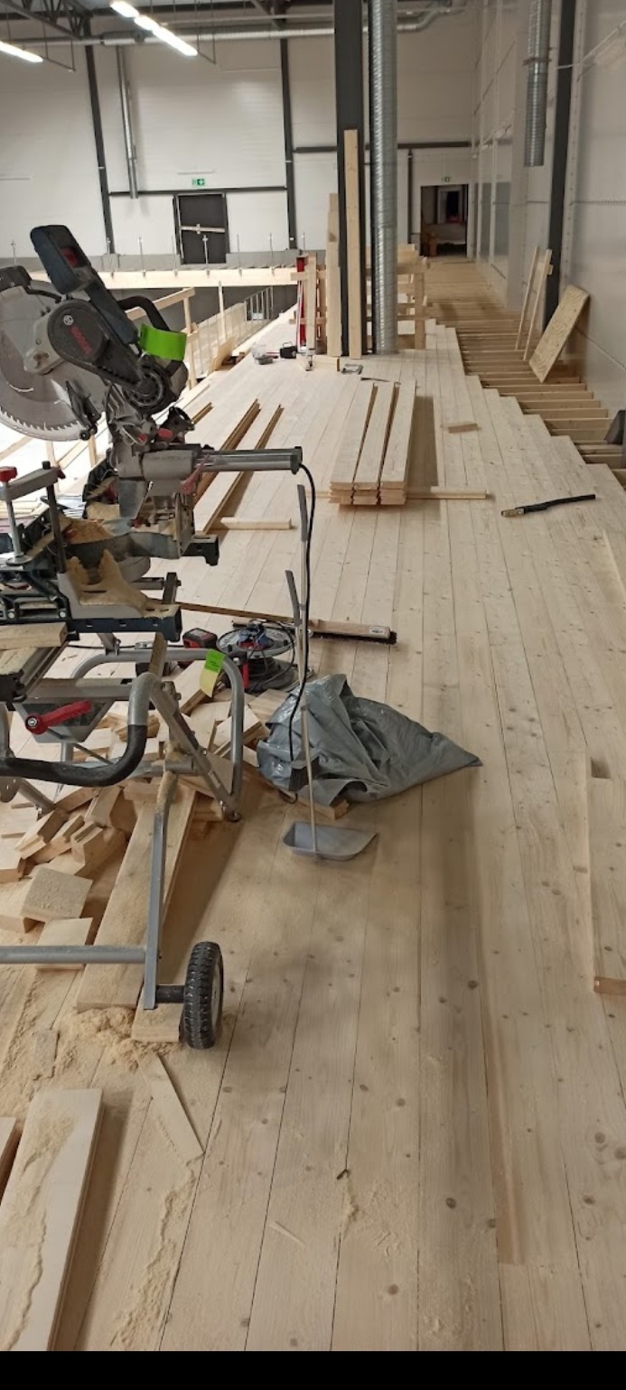 Układanie jasnej drewnianej podłogi w przestronnym wnętrzu, widoczna piła ukosowa i stosy desek, w tle metalowa konstrukcja antresoli.