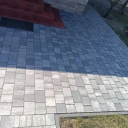 Nowo ułożona, szara kostka brukowa prowadząca do schodów wejściowych, z widoczną wycieraczką i fragmentem trawnika po prawej stronie.