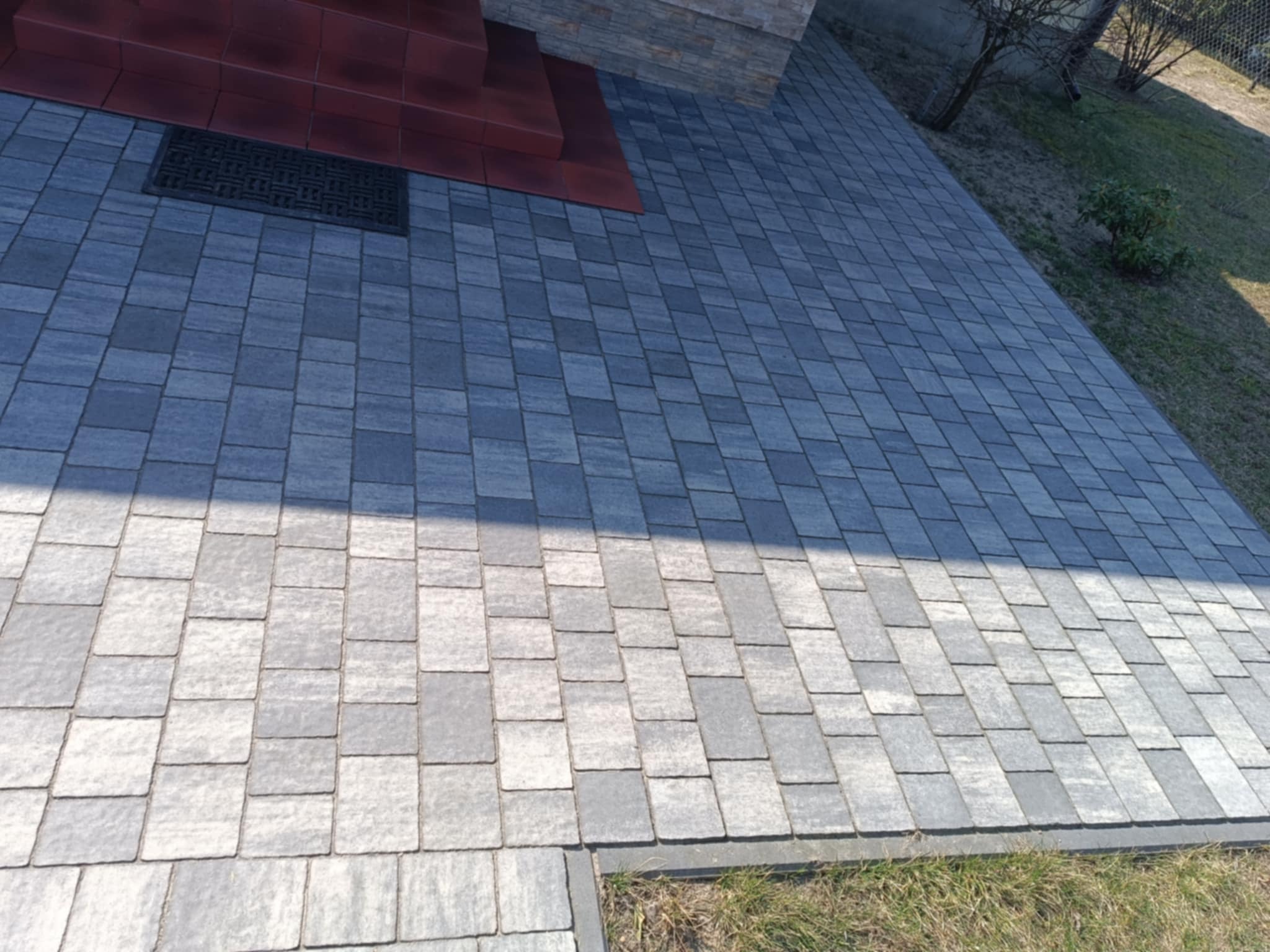 Nowo ułożona, szara kostka brukowa prowadząca do schodów wejściowych, z widoczną wycieraczką i fragmentem trawnika po prawej stronie.