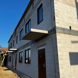 InstalBud DD - Surowy budynek z betonu komórkowego w trakcie budowy, widoczne okna, drzwi i instalacja elektryczna na elewacji. Niebo błękitne, słoneczny dzień.