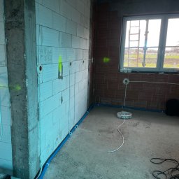 InstalBud DD - Wnętrze w trakcie prac elektrycznych: kable w peszlach przy ścianie z betonu komórkowego, puszki instalacyjne, okno z widokiem na zewnątrz. Etap prac instalacyjnych.
