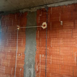 InstalBud DD - Czerwona ściana z pustaków z instalacją elektryczną w trakcie montażu: kable w peszlach, puszki podtynkowe. Widoczne ślady zaprawy i betonu.