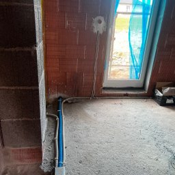 InstalBud DD - Instalacja elektryczna w nowym budynku: kable w peszlach prowadzone wzdłuż ściany z cegły, puszka elektryczna pod oknem. Widoczne surowe ściany i betonowa podłoga.