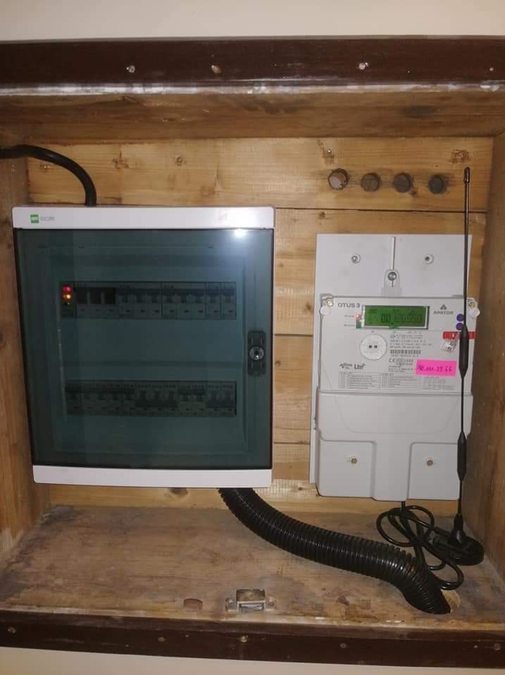 Zamknięta skrzynka bezpiecznikowa z licznikiem energii elektrycznej w drewnianej obudowie. Widoczne przewody i elementy instalacji.