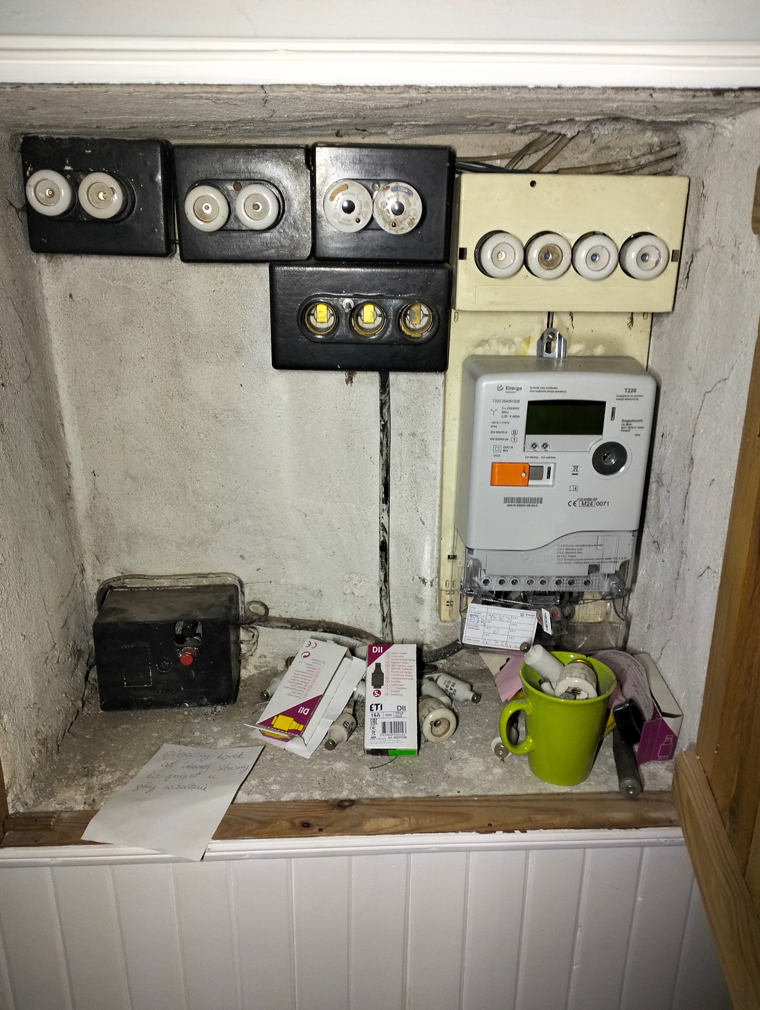 Wnęka z instalacją elektryczną: stare bezpieczniki topikowe w czarnych i kremowych obudowach, licznik energii i zielony kubek z bezpiecznikami na półce.