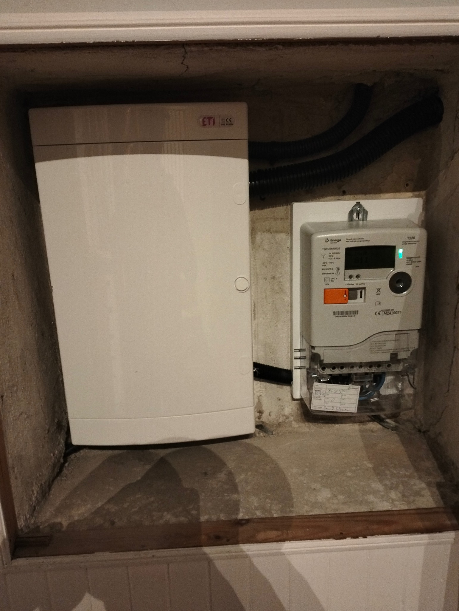 Zamknięta skrzynka elektryczna i licznik energii elektrycznej wnęce. Widoczne kable w peszlu. Szare tło ściany i podłogi.