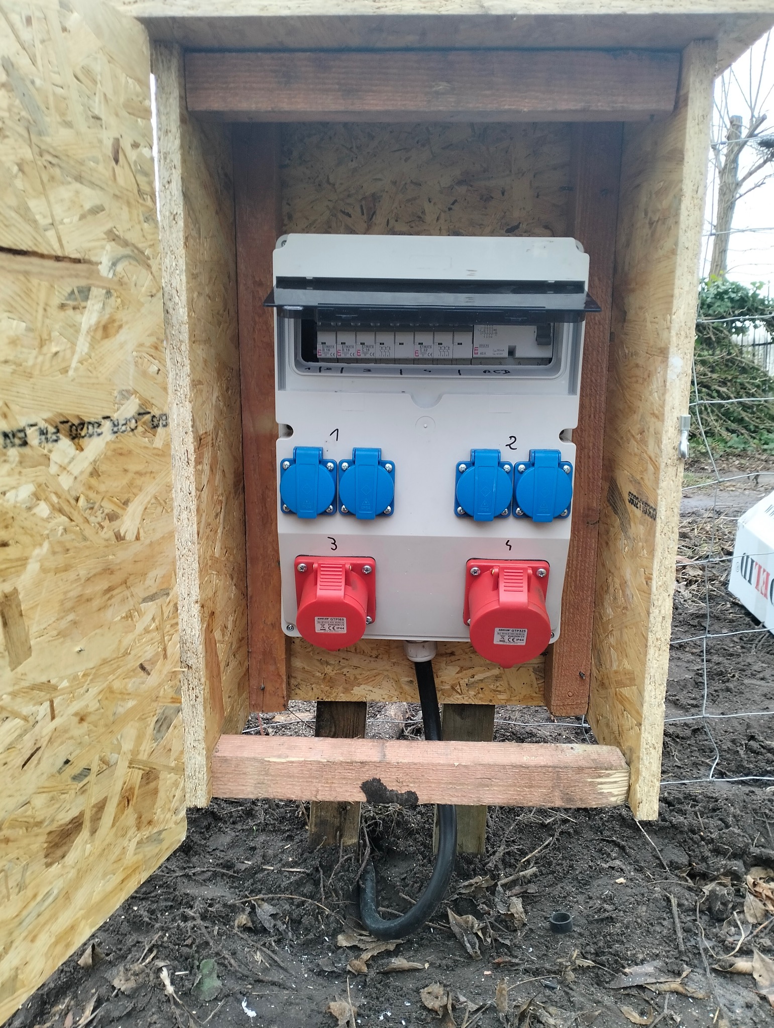 Skrzynka elektryczna z bezpiecznikami i gniazdami (2 niebieskie, 2 czerwone) w drewnianej obudowie na zewnątrz, widoczne przewody.