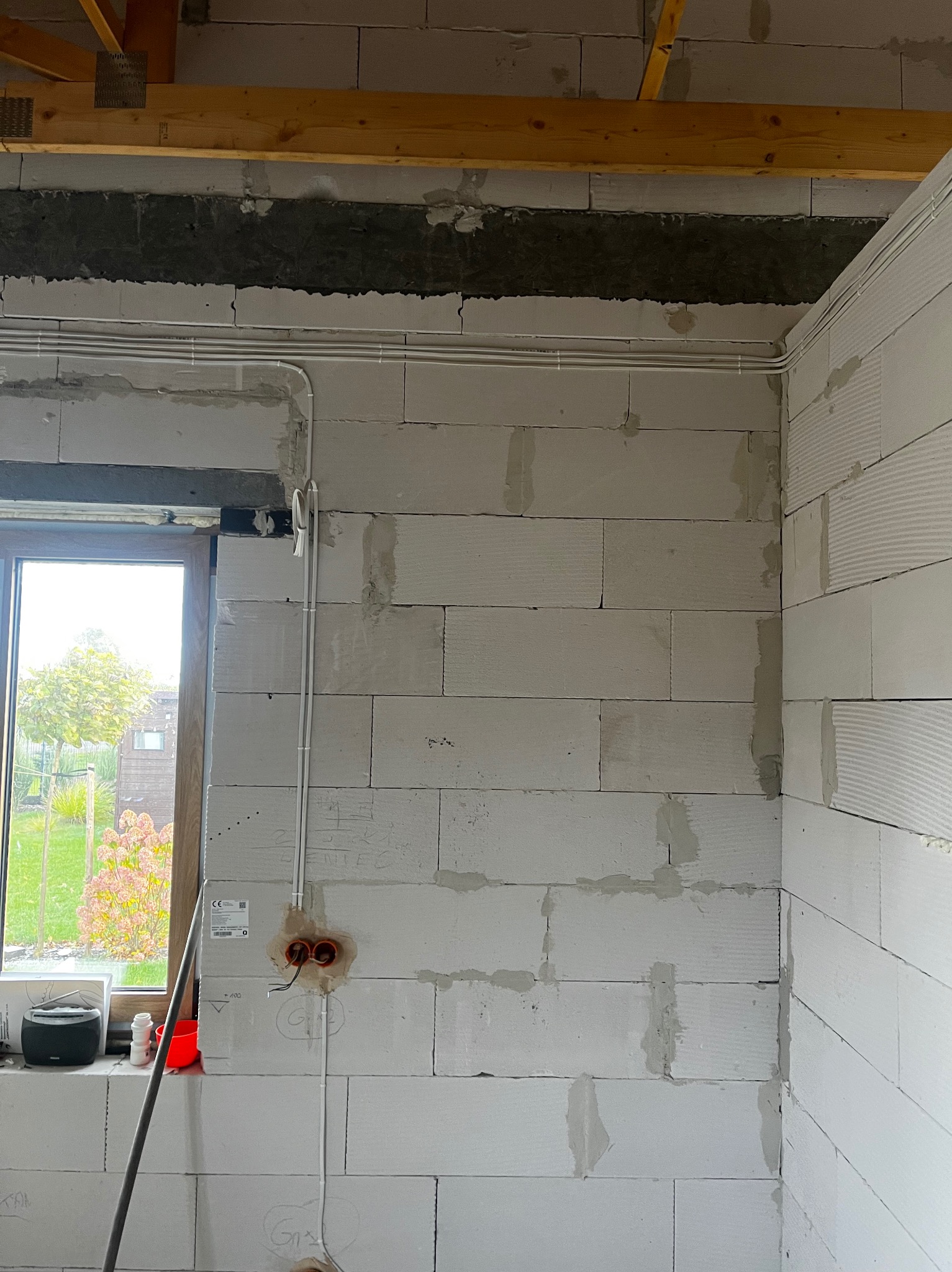 Białe ściany z betonu komórkowego w trakcie montażu instalacji elektrycznej. Widoczne rury instalacyjne, puszki elektryczne i okno z widokiem na zewnątrz.