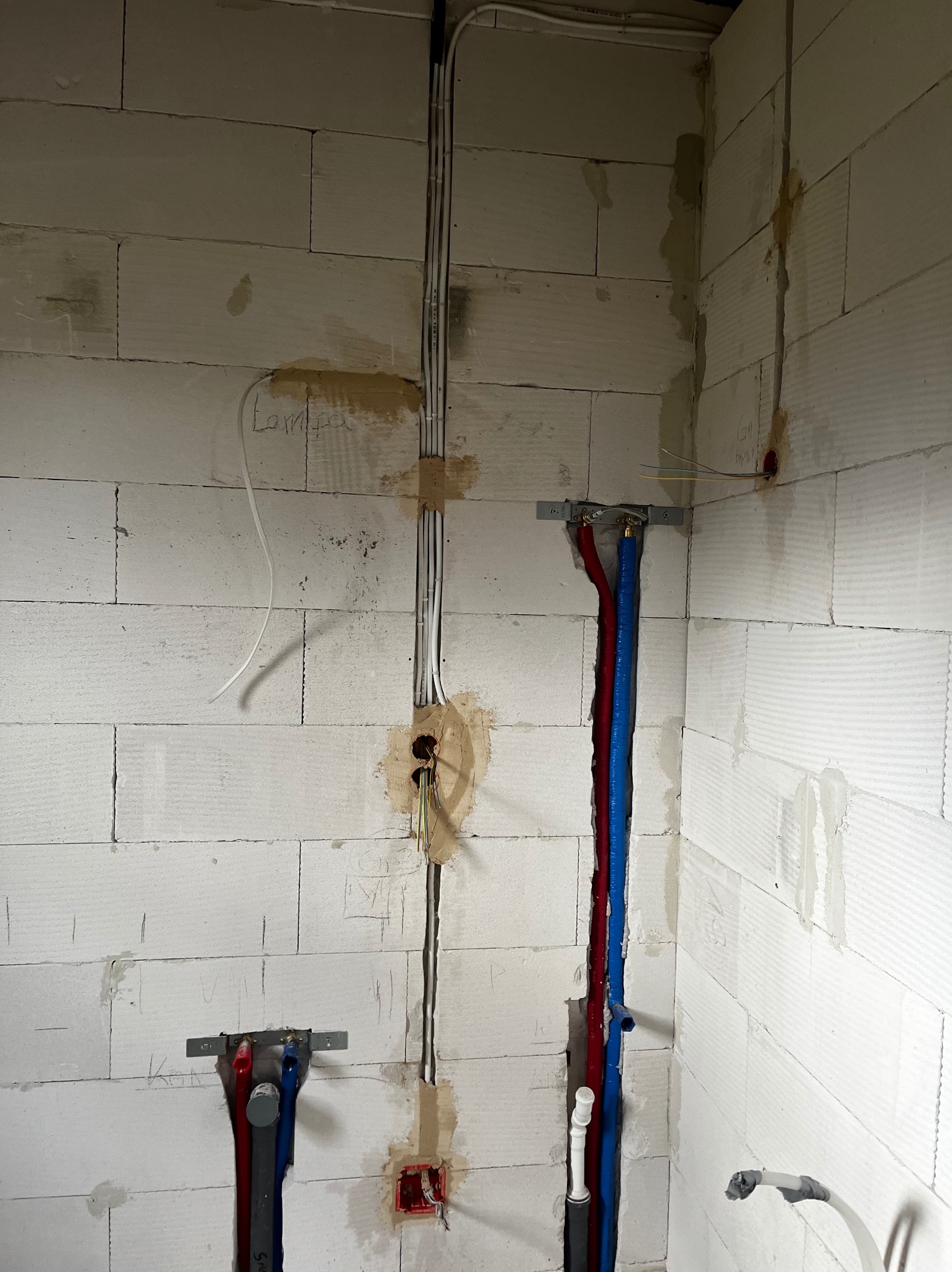 Ściana z betonu komórkowego z bruzdami, w których poprowadzone są instalacje elektryczne i rury wodne w kolorach niebieskim i czerwonym.