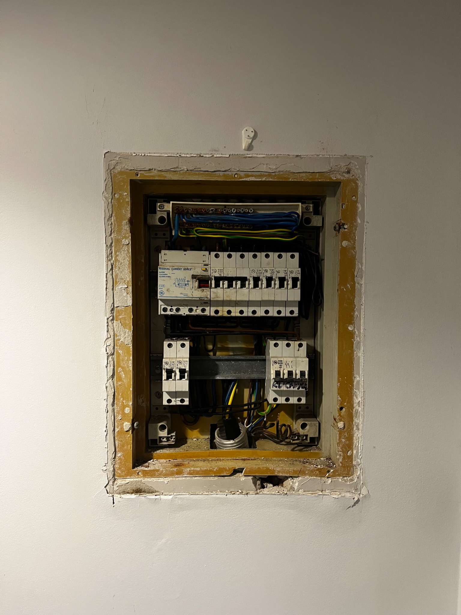 Wnęka w ścianie z zamontowaną skrzynką elektryczną z bezpiecznikami, widoczne przewody i elementy instalacji. Wykończenie wnęki wymaga poprawek.