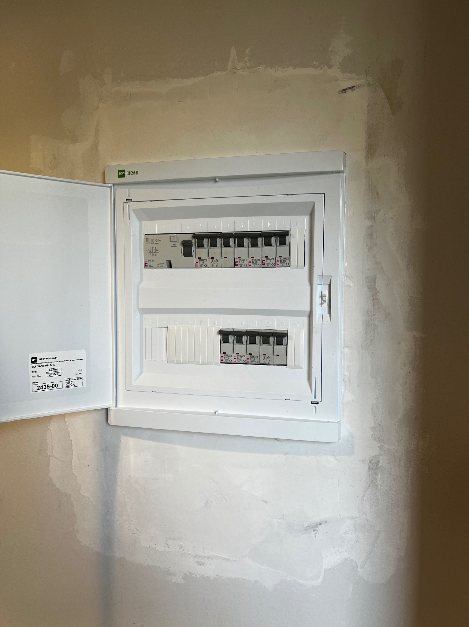 Otwarta, biała skrzynka elektryczna ETI Elegant RP 2x12 z bezpiecznikami na ścianie w trakcie prac wykończeniowych. Widoczne ślady po szpachlowaniu.