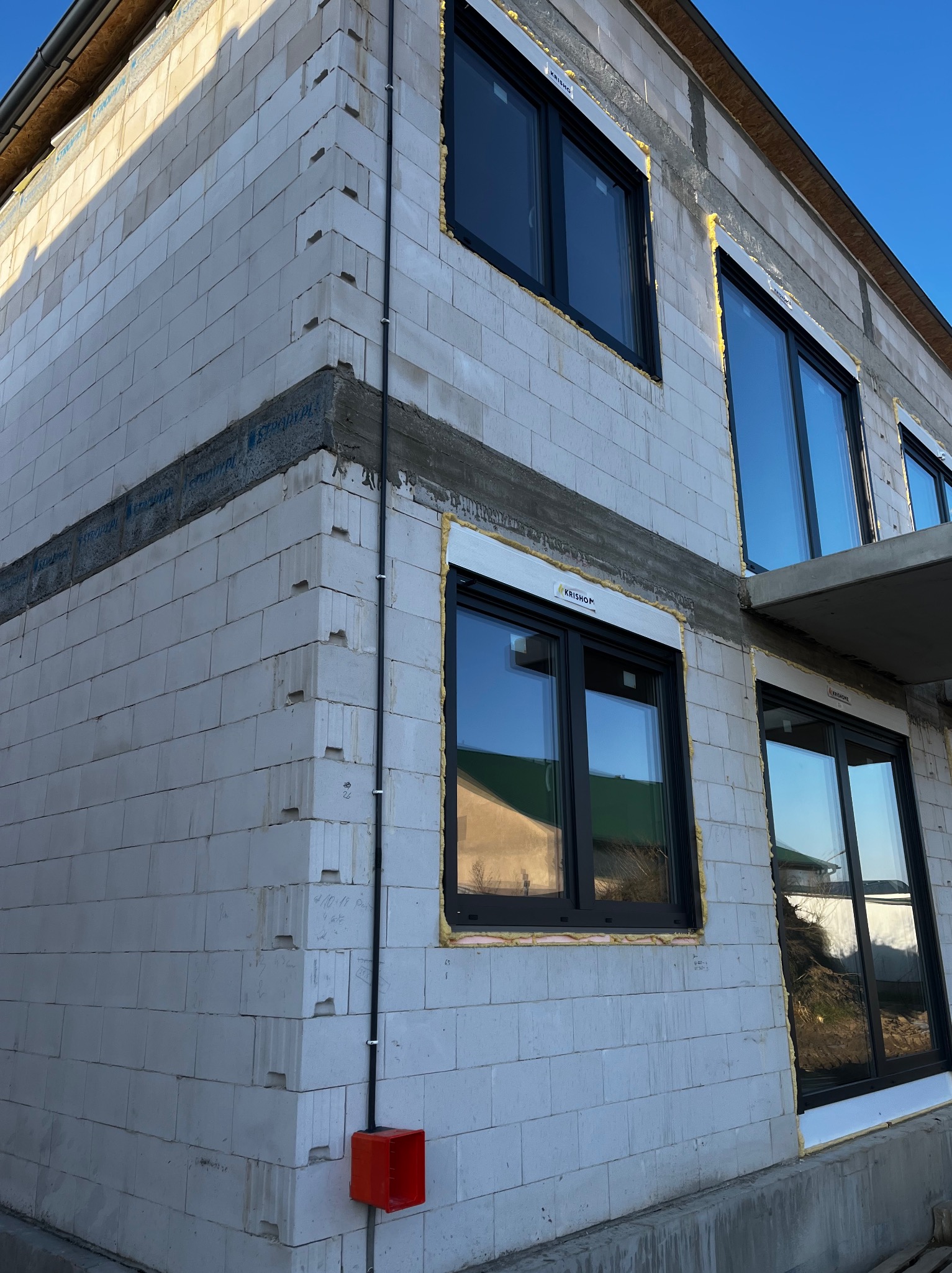 Surowy budynek z betonu komórkowego w trakcie budowy, z zamontowanymi oknami z czarnymi ramami i widoczną instalacją elektryczną w czerwonej puszce.