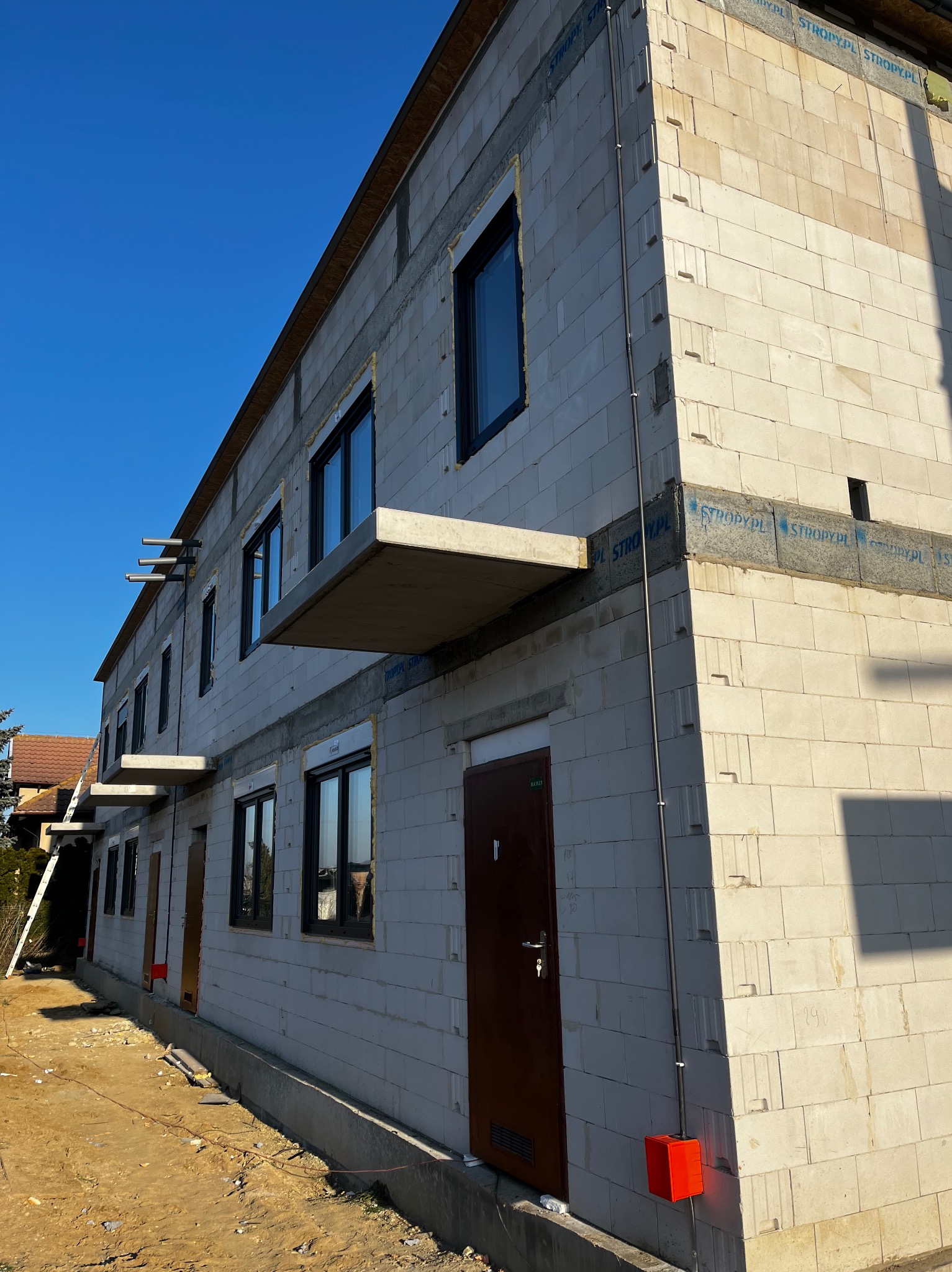 Surowy budynek z betonu komórkowego w trakcie budowy, widoczne okna, drzwi i instalacja elektryczna na elewacji. Niebo błękitne, słoneczny dzień.