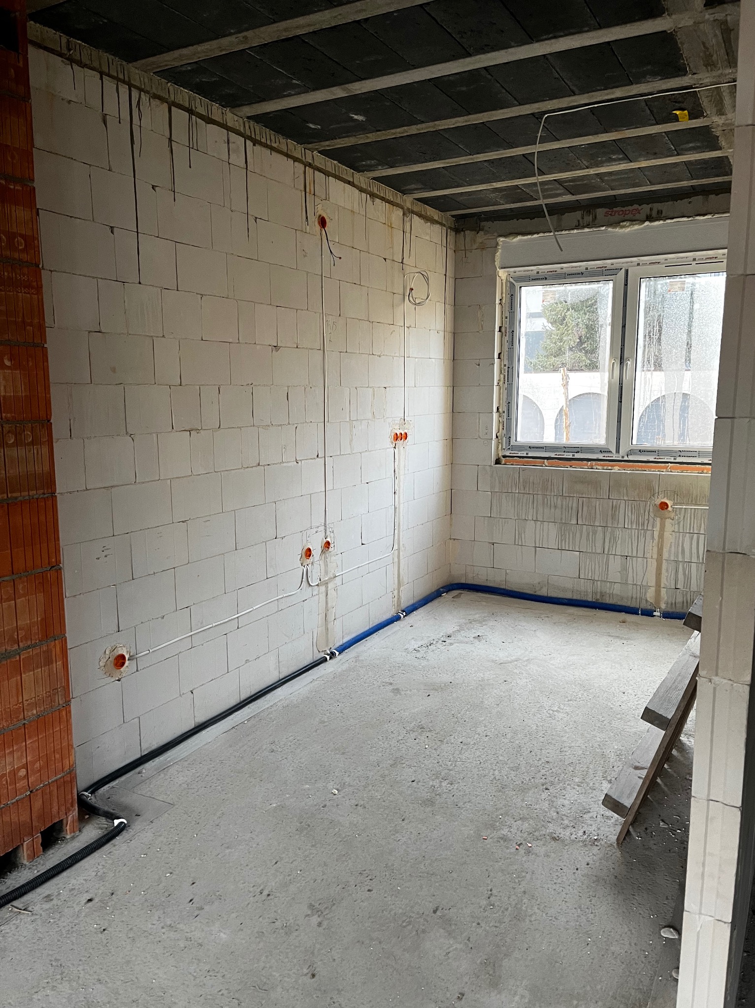 Surowy pokój z instalacją elektryczną i rurami w ścianach z białych bloków. Widoczne puszki elektryczne i okno z widokiem na zewnątrz. Podłoga betonowa, sufit z płyt.