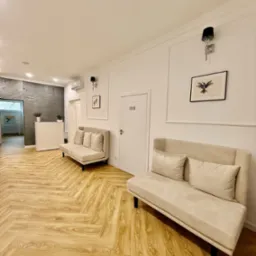 Jasny, przestronny hol z dwiema beżowymi sofami, drewnianą podłogą ułożoną w jodełkę, recepcją w tle i minimalistycznymi dekoracjami ściennymi.