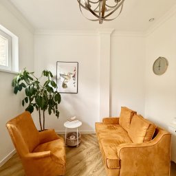 CENTRUM WSPARCIA DOBROSTAN SPÓŁKA Z OGRANICZONĄ ODPOWIEDZIALNOŚCIĄ - Jasne wnętrze z pomarańczowym fotelem i kanapą, rośliną doniczkową, obrazkiem kolibra i zegarem na ścianie.