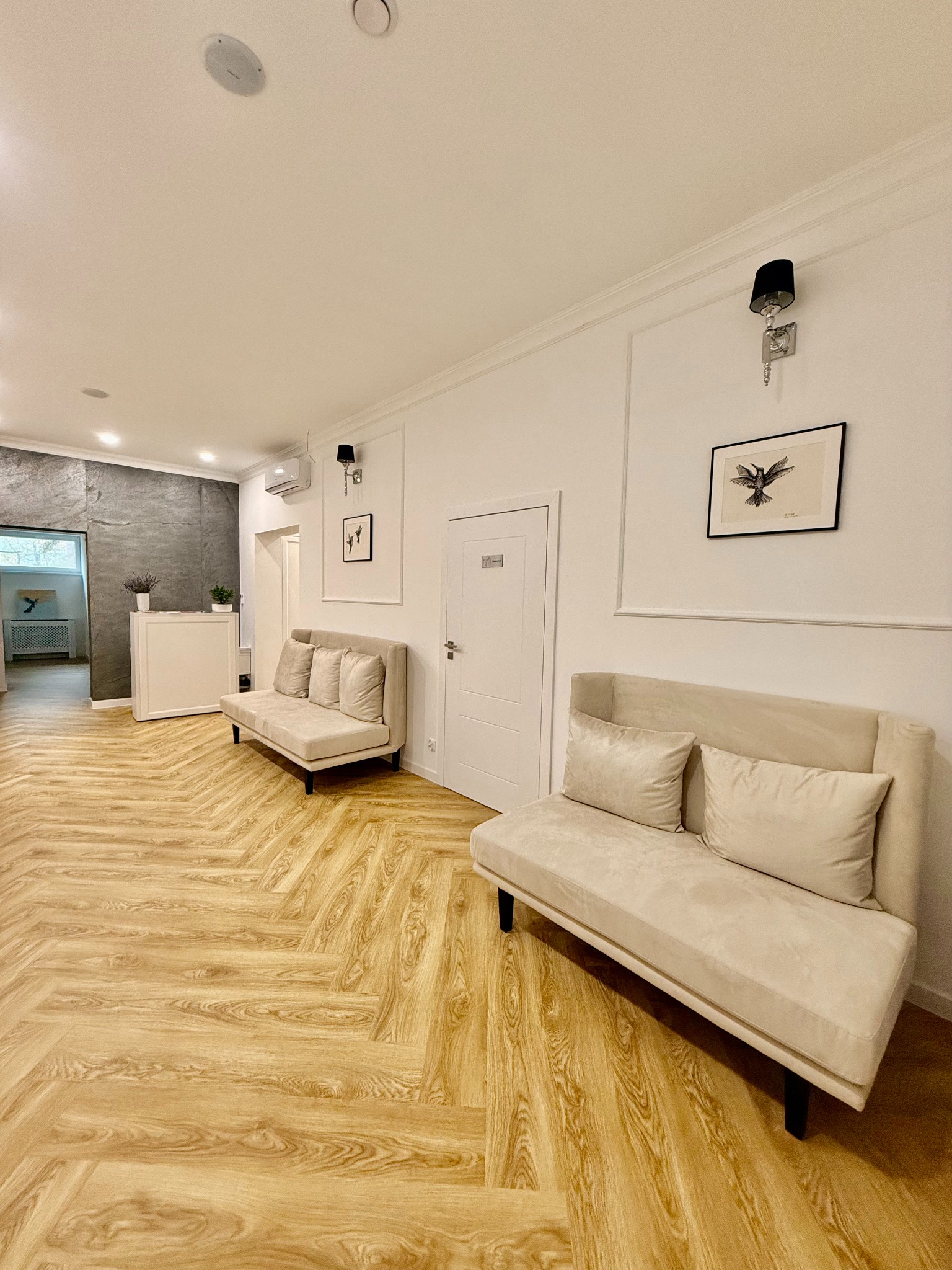 Jasny, przestronny hol z dwiema beżowymi sofami, drewnianą podłogą ułożoną w jodełkę, recepcją w tle i minimalistycznymi dekoracjami ściennymi.