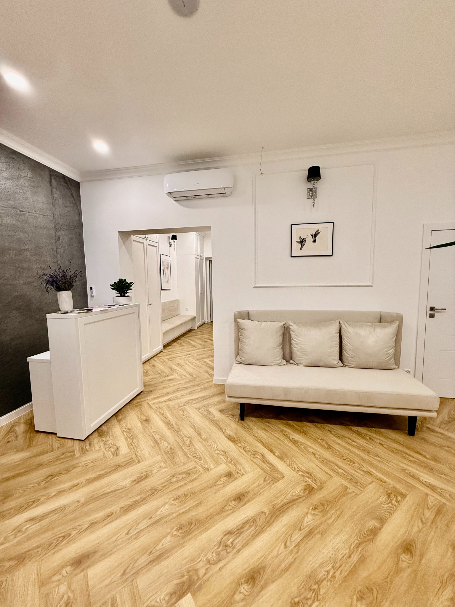 Jasne wnętrze poczekalni z recepcją, sofą z poduszkami i wzorzystą podłogą z paneli, widok na korytarz z drzwiami i schodami.