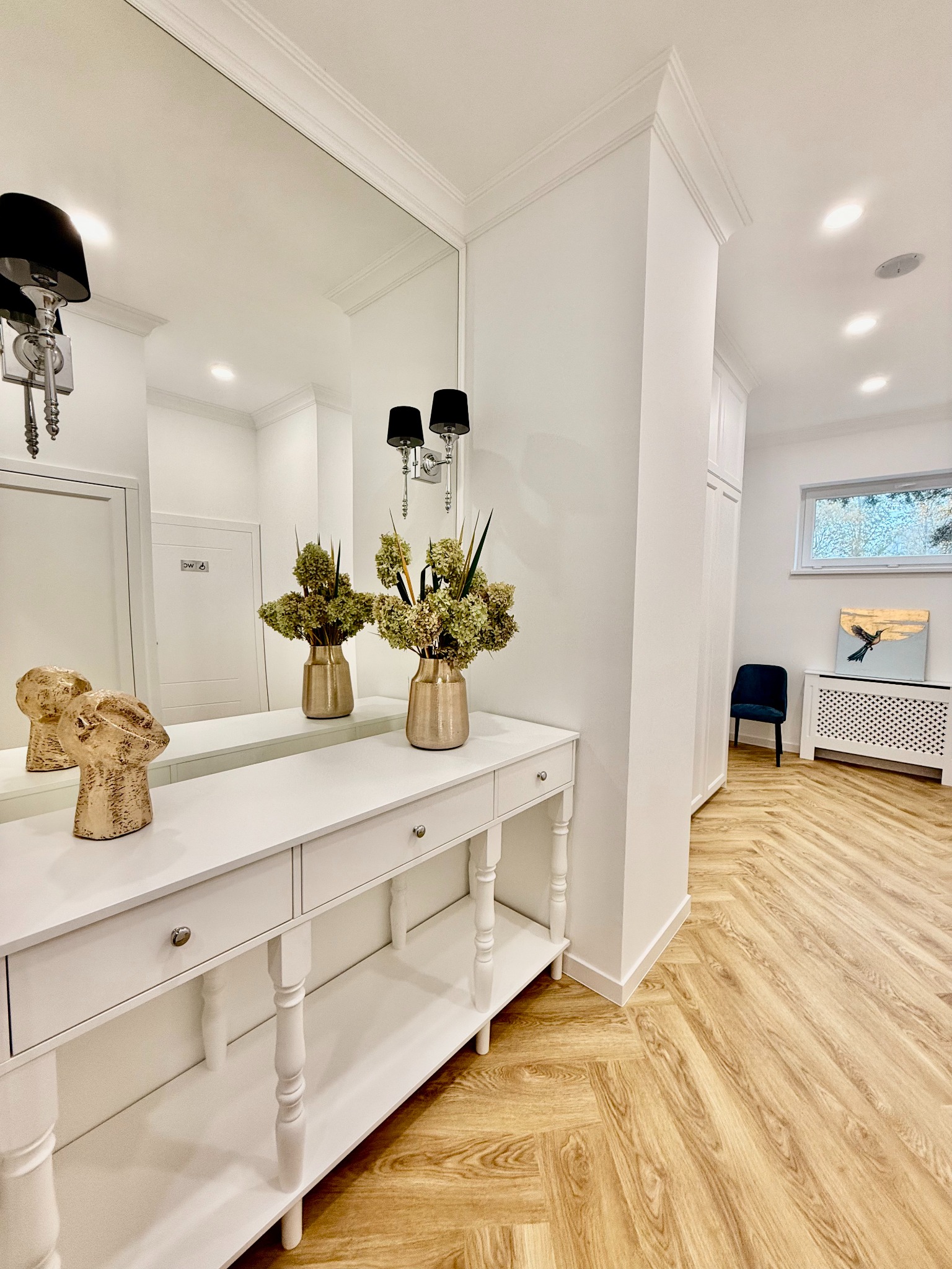 Jasny, elegancki przedpokój z parkietem w jodełkę, białą konsolą z lustrem, złotymi wazonami z suchymi kwiatami i rzeźbą głowy; w tle widoczny pokój z granatowym krzesłem i obrazem ptaka.