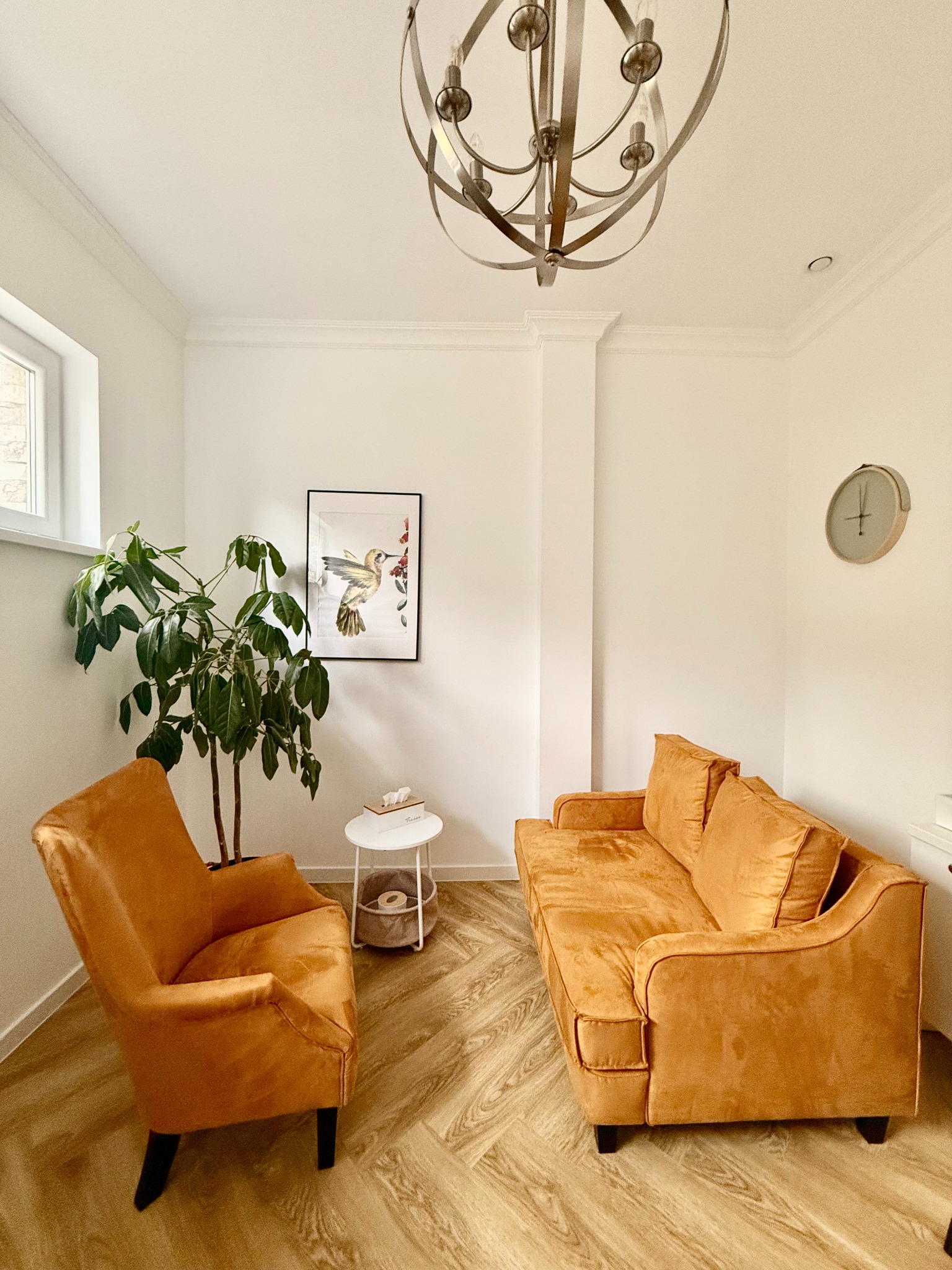 Jasne wnętrze z pomarańczowym fotelem i kanapą, rośliną doniczkową, obrazkiem kolibra i zegarem na ścianie.