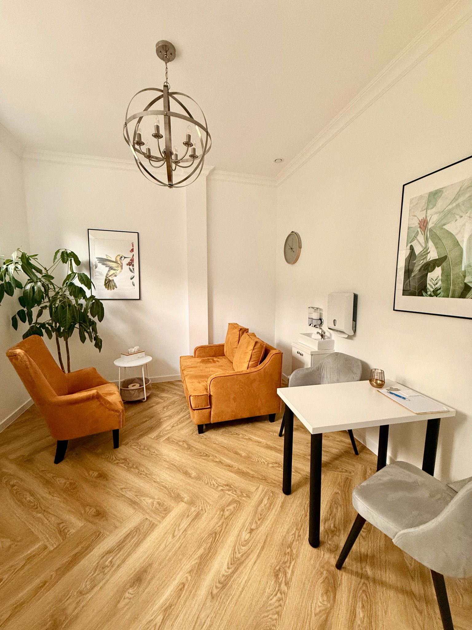 Jasne wnętrze gabinetu z pomarańczowymi fotelami i sofą, drewnianą podłogą w jodełkę, rośliną doniczkową i minimalistycznymi obrazami na ścianach, sugerujące profesjonalne otoczenie terapeutyczne.