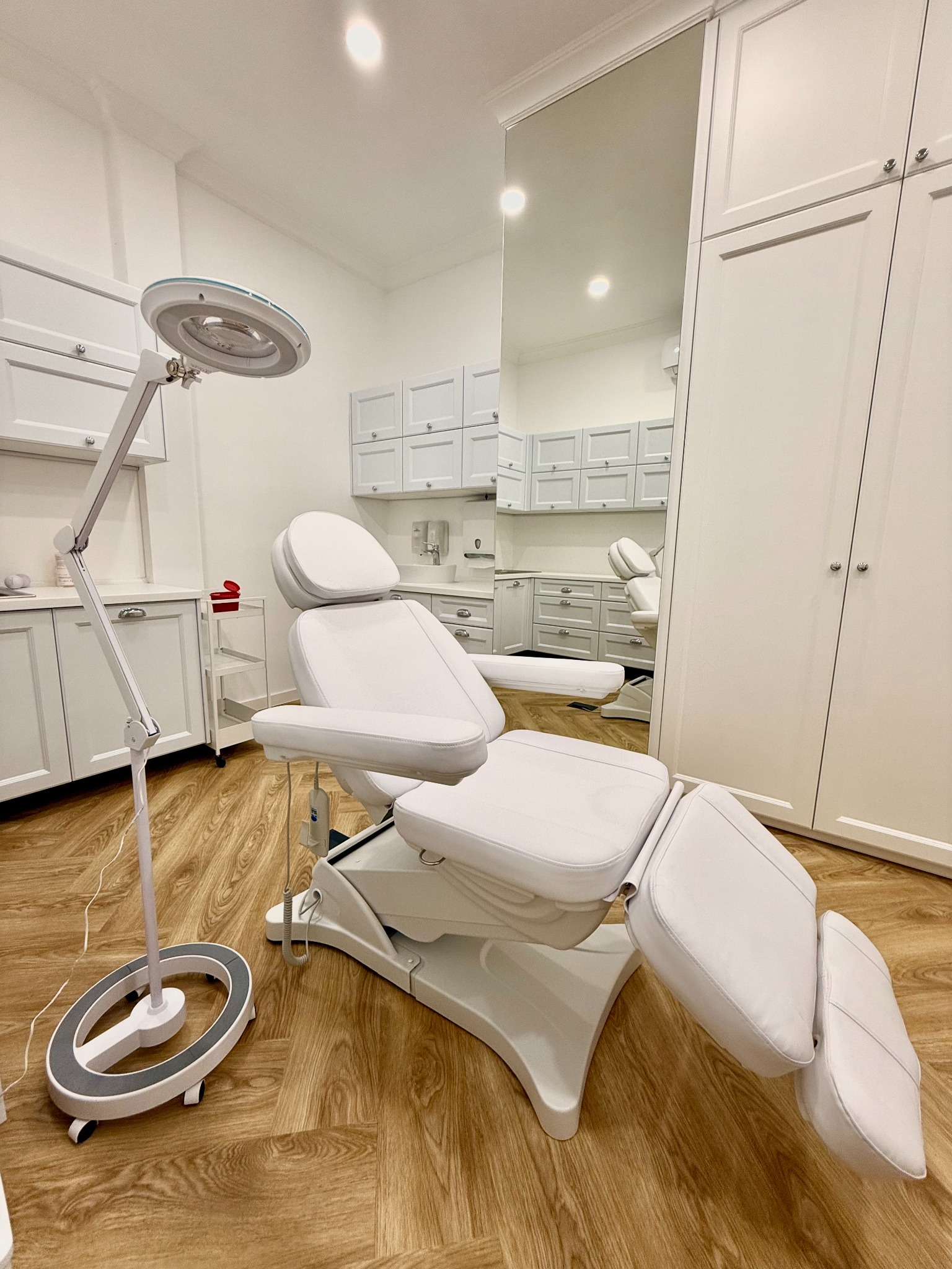 Biały, regulowany fotel zabiegowy z lampą powiększającą w jasnym gabinecie z drewnianą podłogą. W tle białe szafki i umywalka.