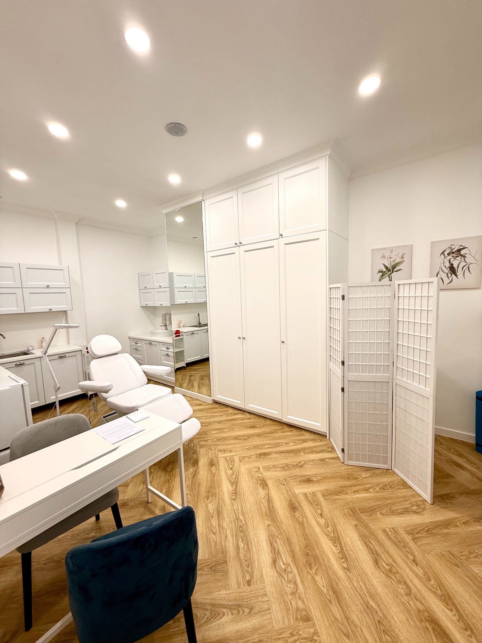 Jasne, przestronne wnętrze gabinetu z fotelem zabiegowym, biurkiem z dokumentami, parawanem i szafkami w tle, oświetlone punktowymi lampami sufitowymi i z podłogą ułożoną w jodełkę.