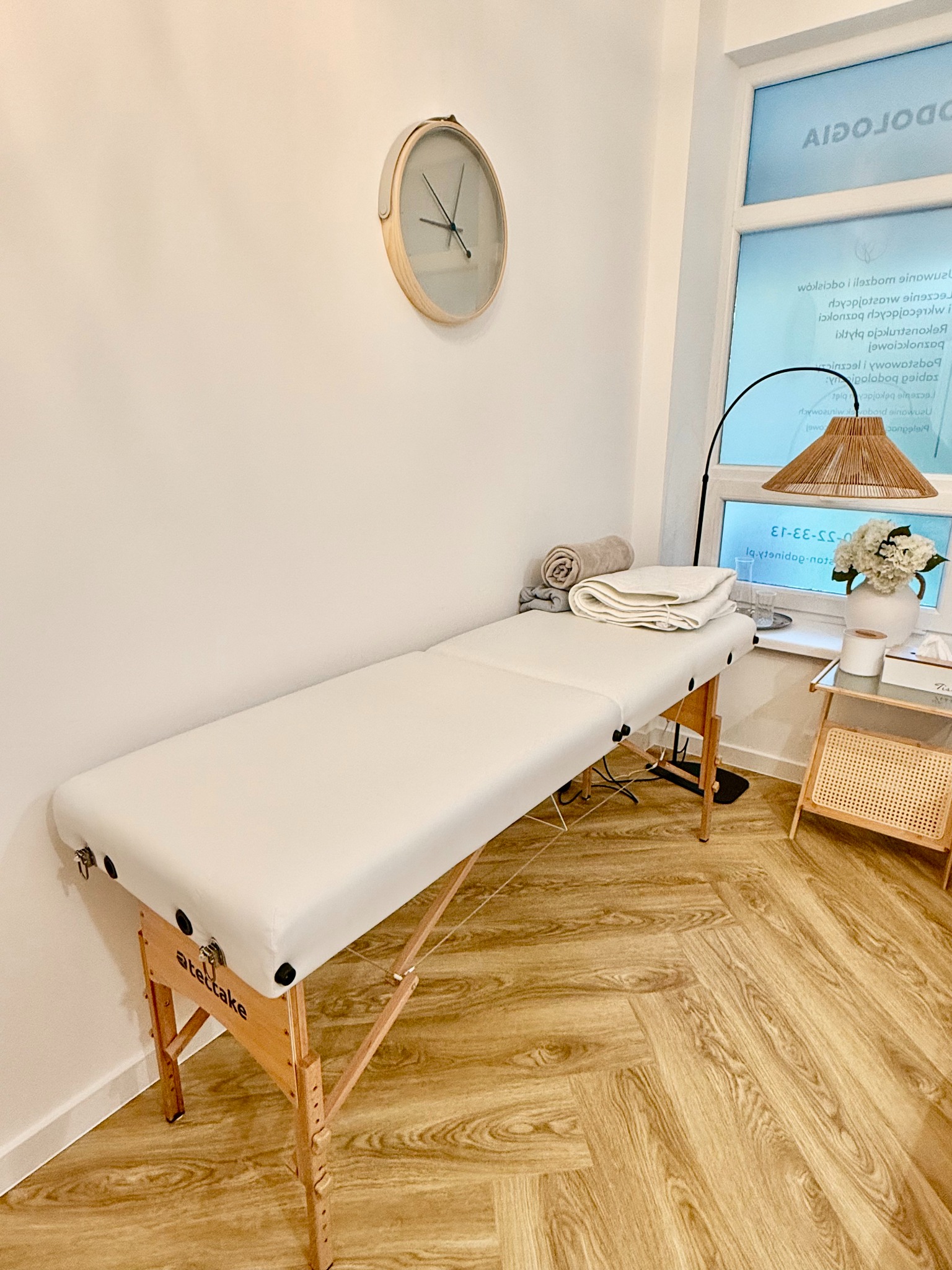 Jasne wnętrze gabinetu z drewnianą podłogą w jodełkę; biały stół do masażu marki Atettake z ręcznikami, zegar na ścianie, lampa z abażurem i stolik z kwiatami w wazonie.