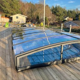 4U SOLUTION SPÓŁKA Z OGRANICZONĄ ODPOWIEDZIALNOŚCIĄ - Montaż zadaszenia basenowego z poliwęglanu na drewnianym tarasie, widoczny pracownik w odblaskowej kamizelce, narzędzia i worki z materiałami budowlanymi w tle.