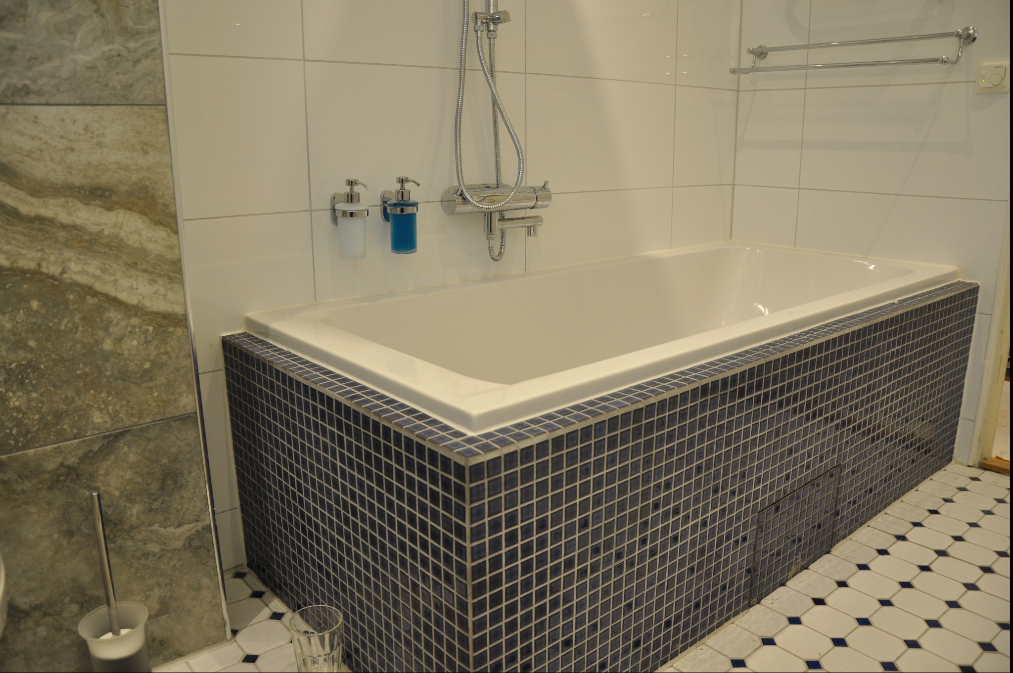 Biała wanna obudowana ciemnymi, mozaikowymi płytkami, z armaturą prysznicową i dozownikami mydła na białych, kwadratowych płytkach ściennych. Na podłodze wzorzyste, biało-niebieskie płytki.