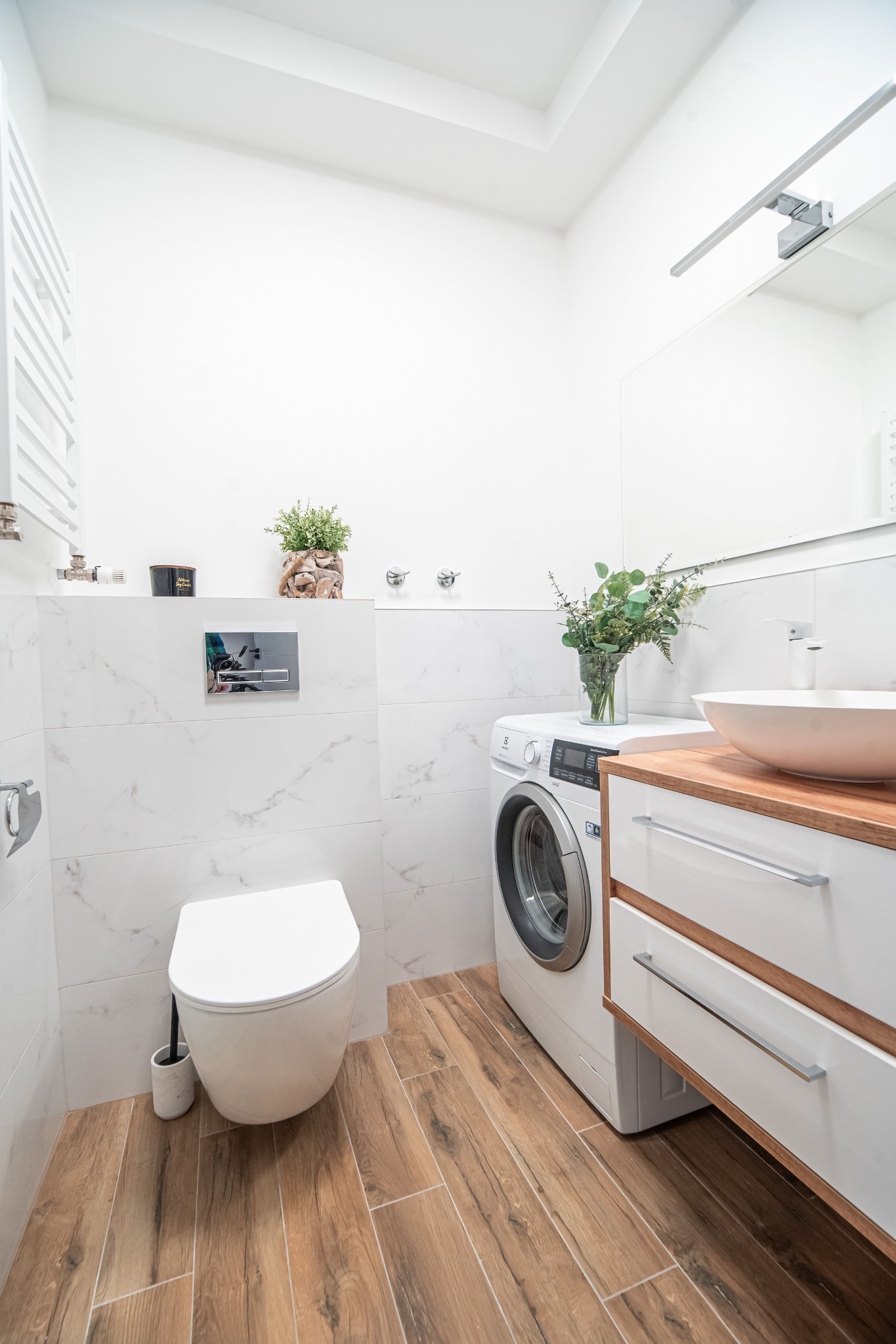 Nowoczesna łazienka z białymi marmurowymi płytkami, drewnopodobną podłogą, wiszącą toaletą, pralką, umywalką nablatową na drewnianym blacie i roślinami dekoracyjnymi.