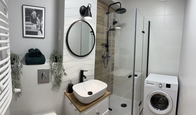 Nowoczesna łazienka z kabiną prysznicową, umywalką nablatową na drewnianym blacie, czarną armaturą, okrągłym lustrem i pralką, dopełniona dekoracyjnymi ręcznikami, roślinami i czarno-białą...