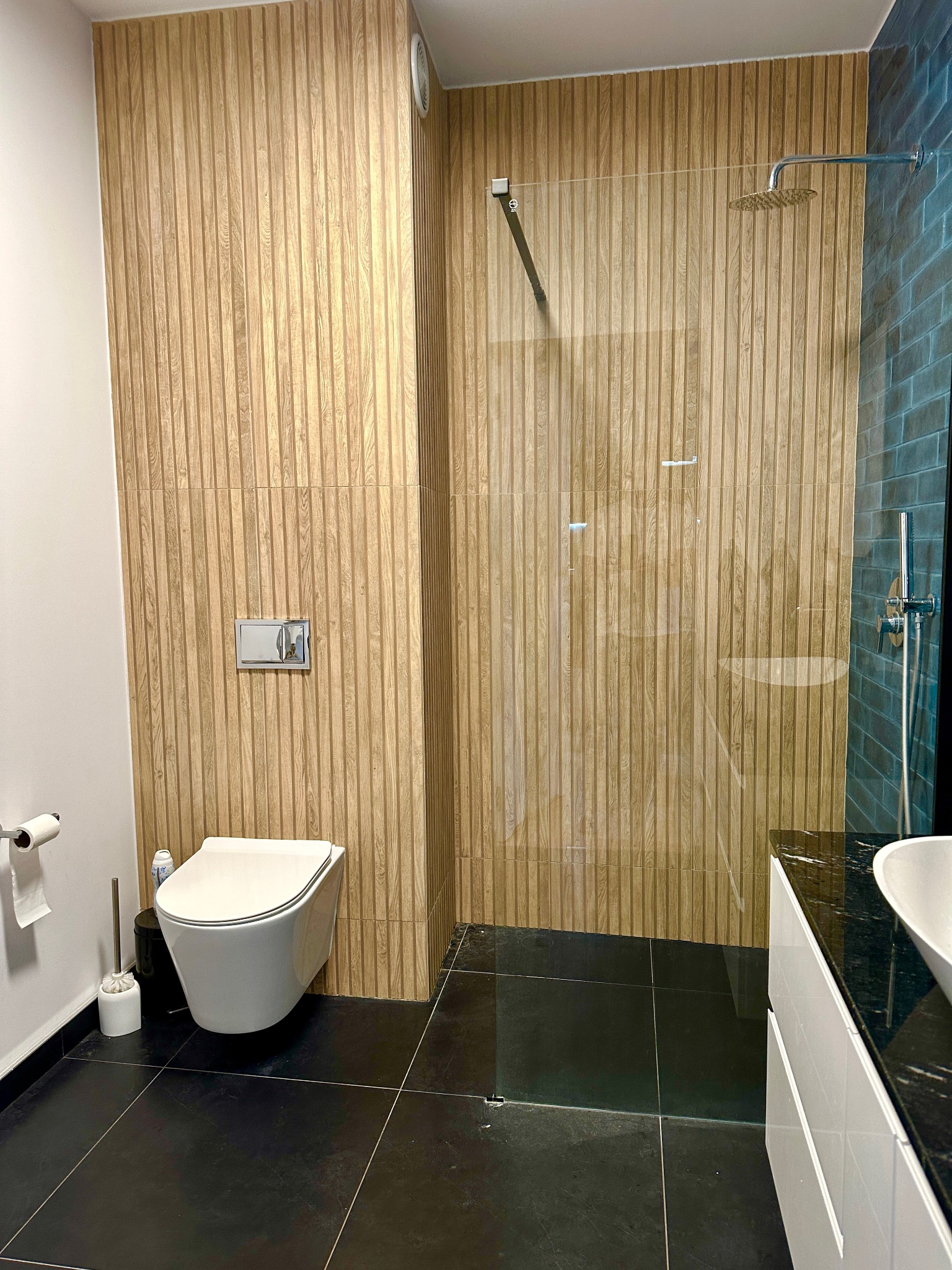 Nowoczesna łazienka z kabiną prysznicową typu walk-in, wykończona płytkami imitującymi drewno i czarną podłogą, z wiszącą miską WC i umywalką z czarnym blatem.