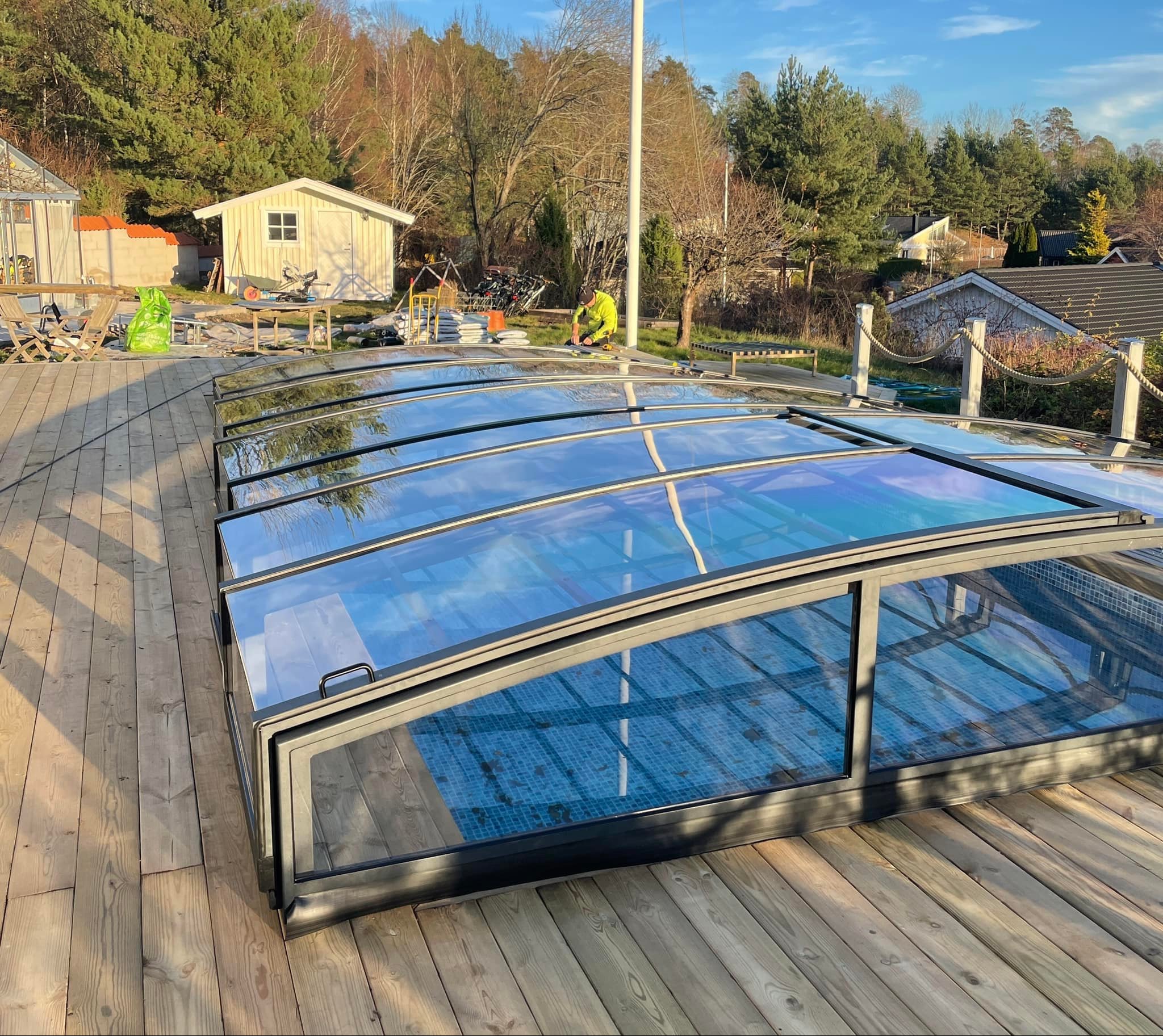 Montaż zadaszenia basenowego z poliwęglanu na drewnianym tarasie, widoczny pracownik w odblaskowej kamizelce, narzędzia i worki z materiałami budowlanymi w tle.