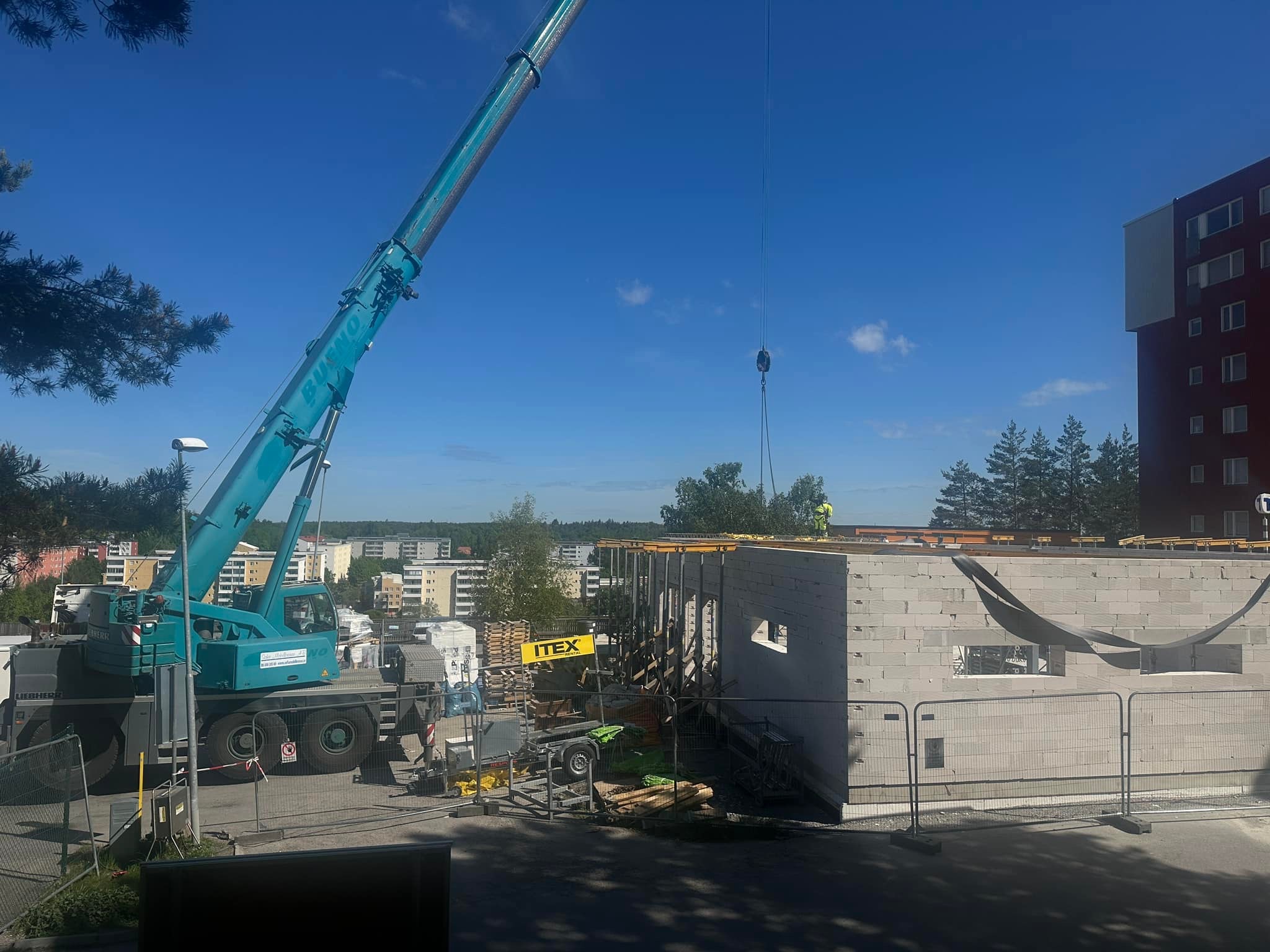 Budowa budynku z bloczków betonowych z użyciem dźwigu, widoczne szalunki i pracownik w żółtym ubraniu na dachu.