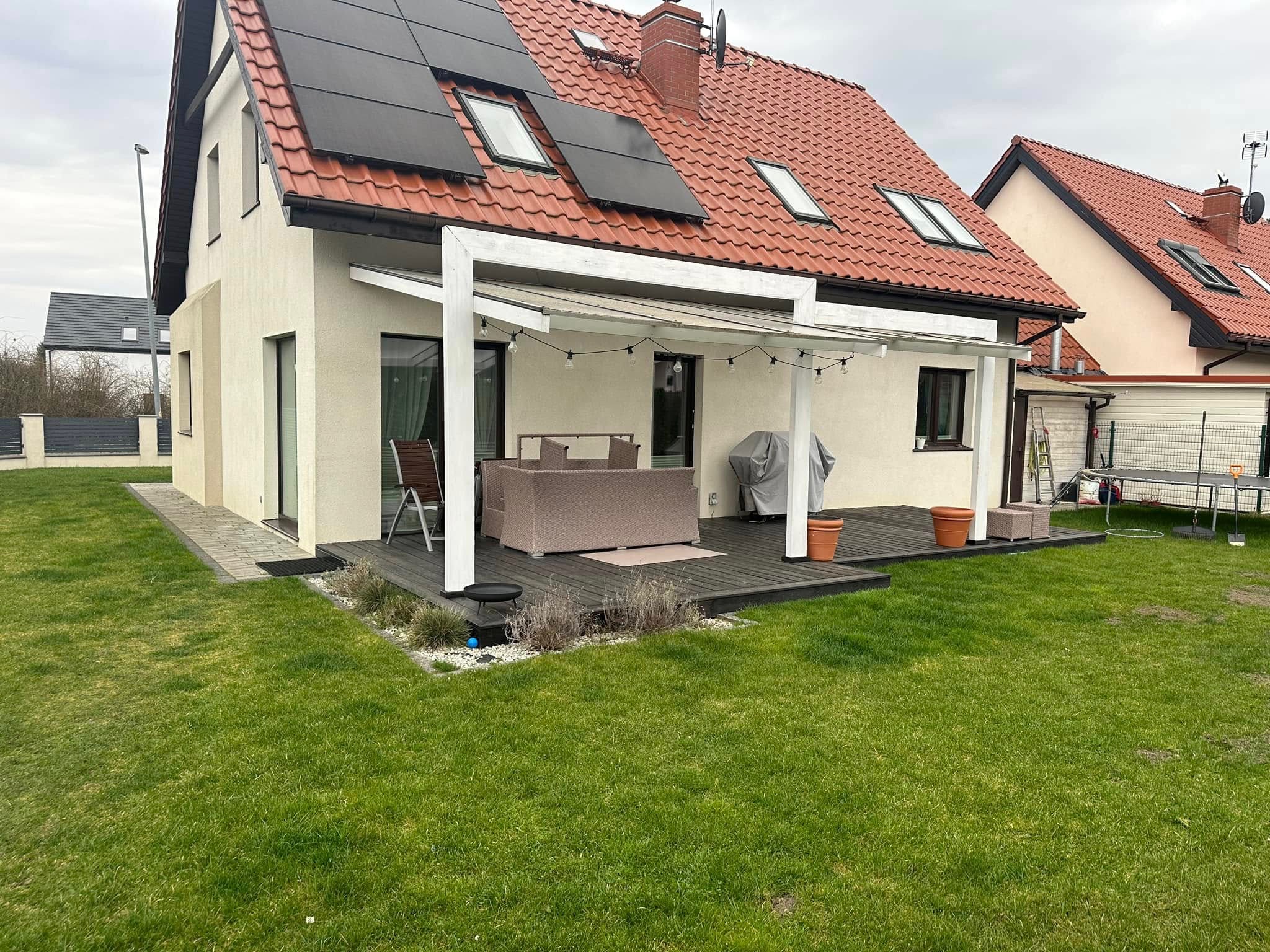 Nowoczesny taras z drewnianą podłogą i białym zadaszeniem, wyposażony w meble ogrodowe z rattanu, grill przy ścianie domu i panele słoneczne na dachu.